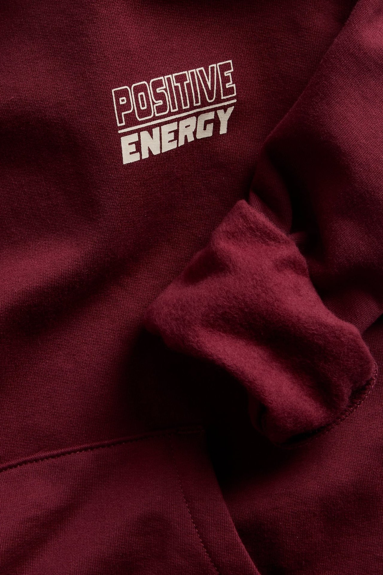 Zbliżenie na bordową bluzę z kapturem z białym napisem „POSITIVE ENERGY” na piersi.