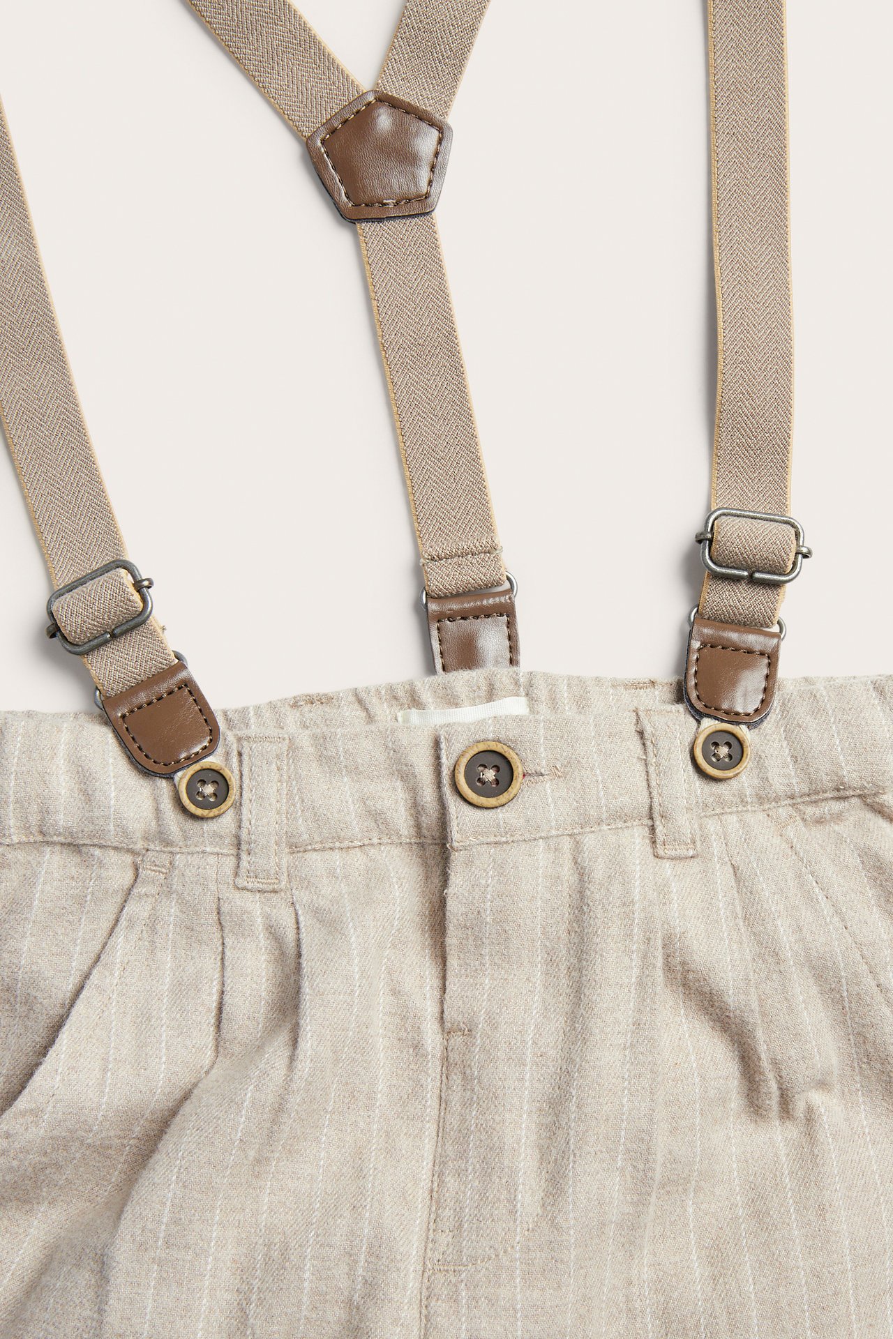 Nærbilde forfra av beige pinstripebukser med brune seler med skinndetaljer.
