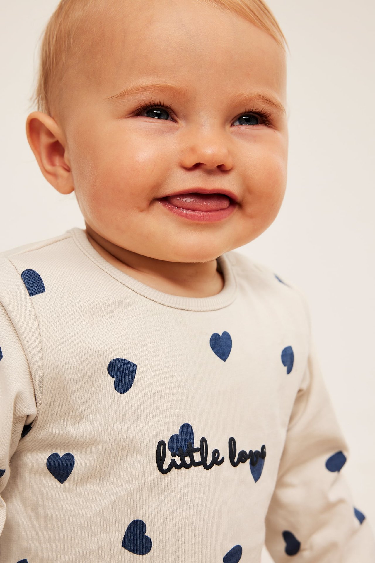 Zbliżenie na dziecko w jasnobeżowej bluzce z długim rękawem z niebieskimi sercami i napisem „little love”.