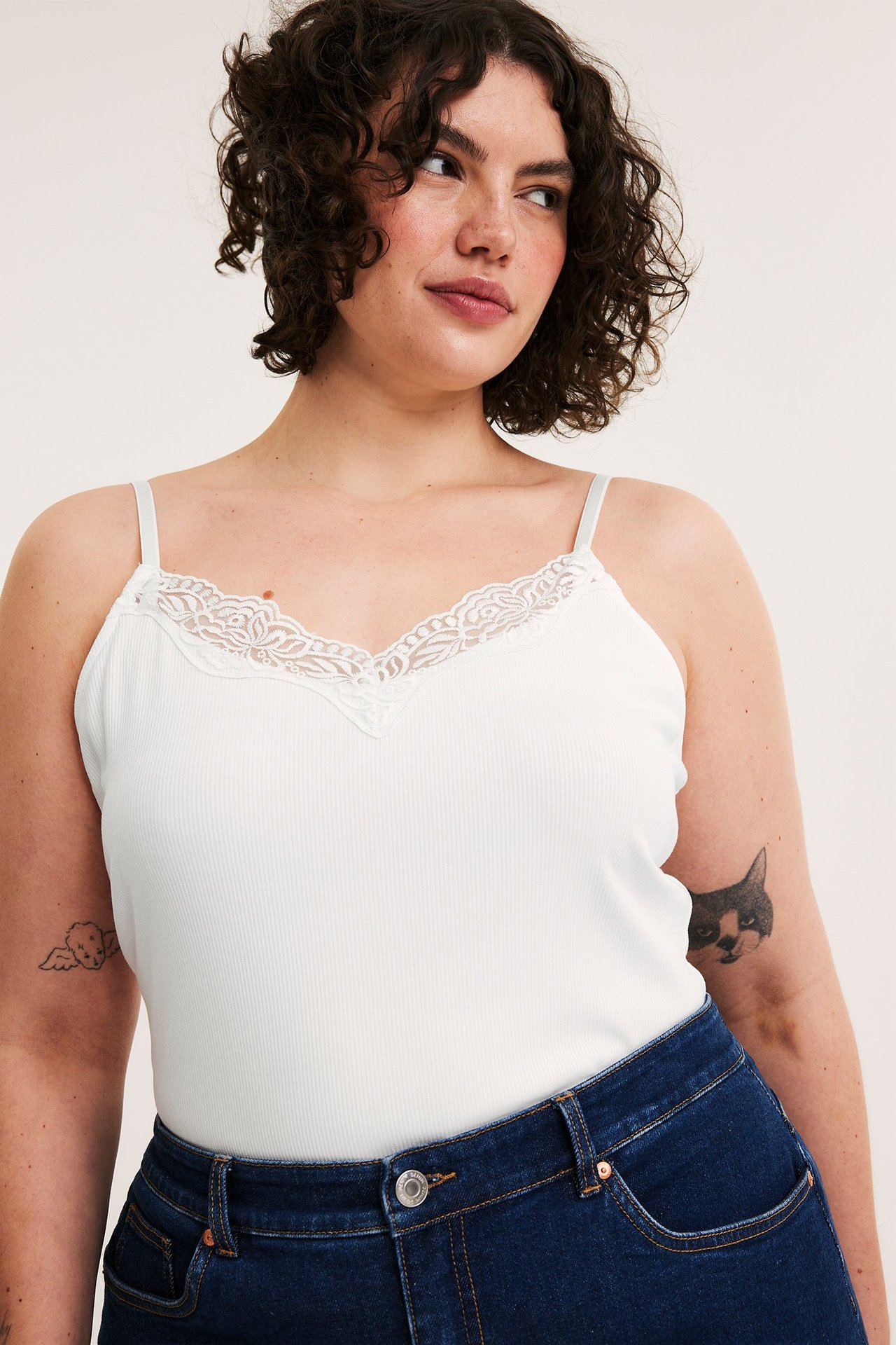 Widok z przodu kobiety w kremowym prążkowanym topie z koronką i ciemnoniebieskich dżinsach.