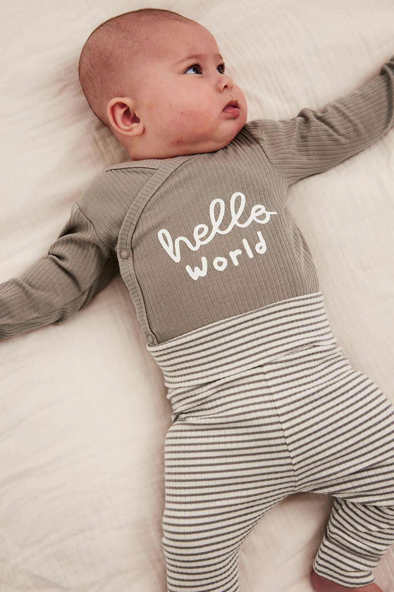 Vauva harmaassa 'hello world' ribbineuleisessa pitkähihaisessa bodyssä ja raidallisissa housuissa, ylhäältä.