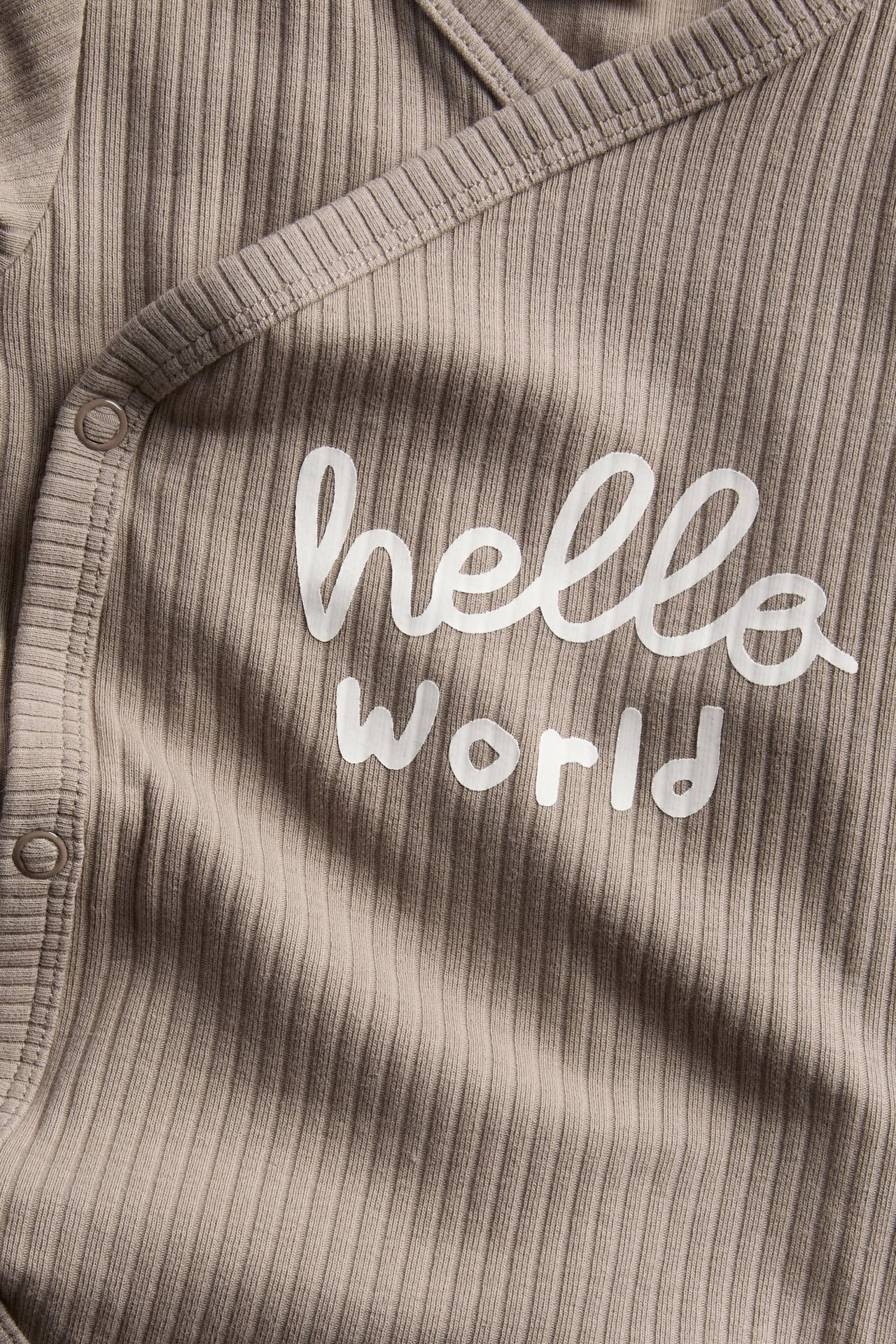 Zbliżenie na jasnobrązowe, prążkowane body niemowlęce z białym napisem 'hello world' i zatrzaskami.