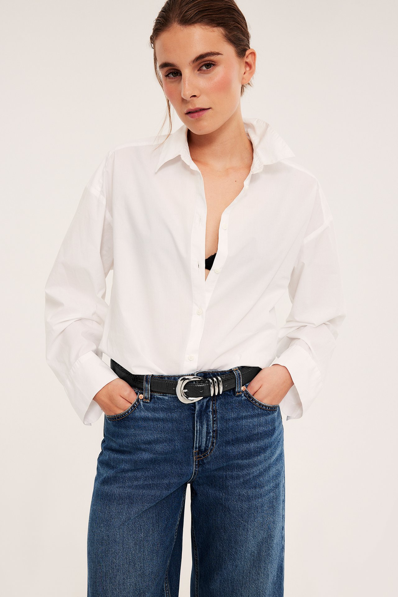 Frontbilde av en kvinne i en hvit langermet skjorte, blå jeans og et svart skinnbelte.