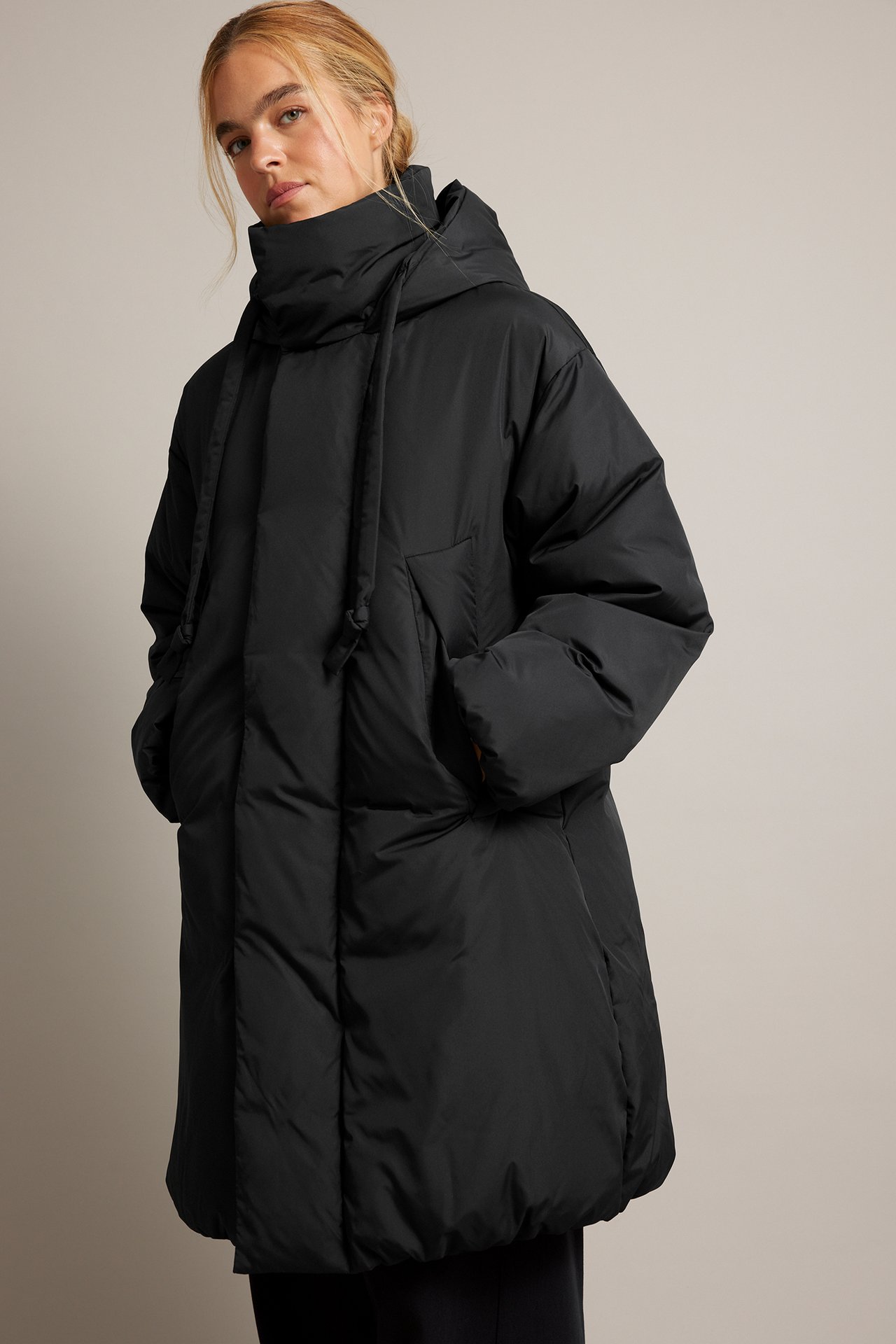 Framifrån, kvinnlig modell i en svart oversized dunjacka med huva och händerna i fickorna.