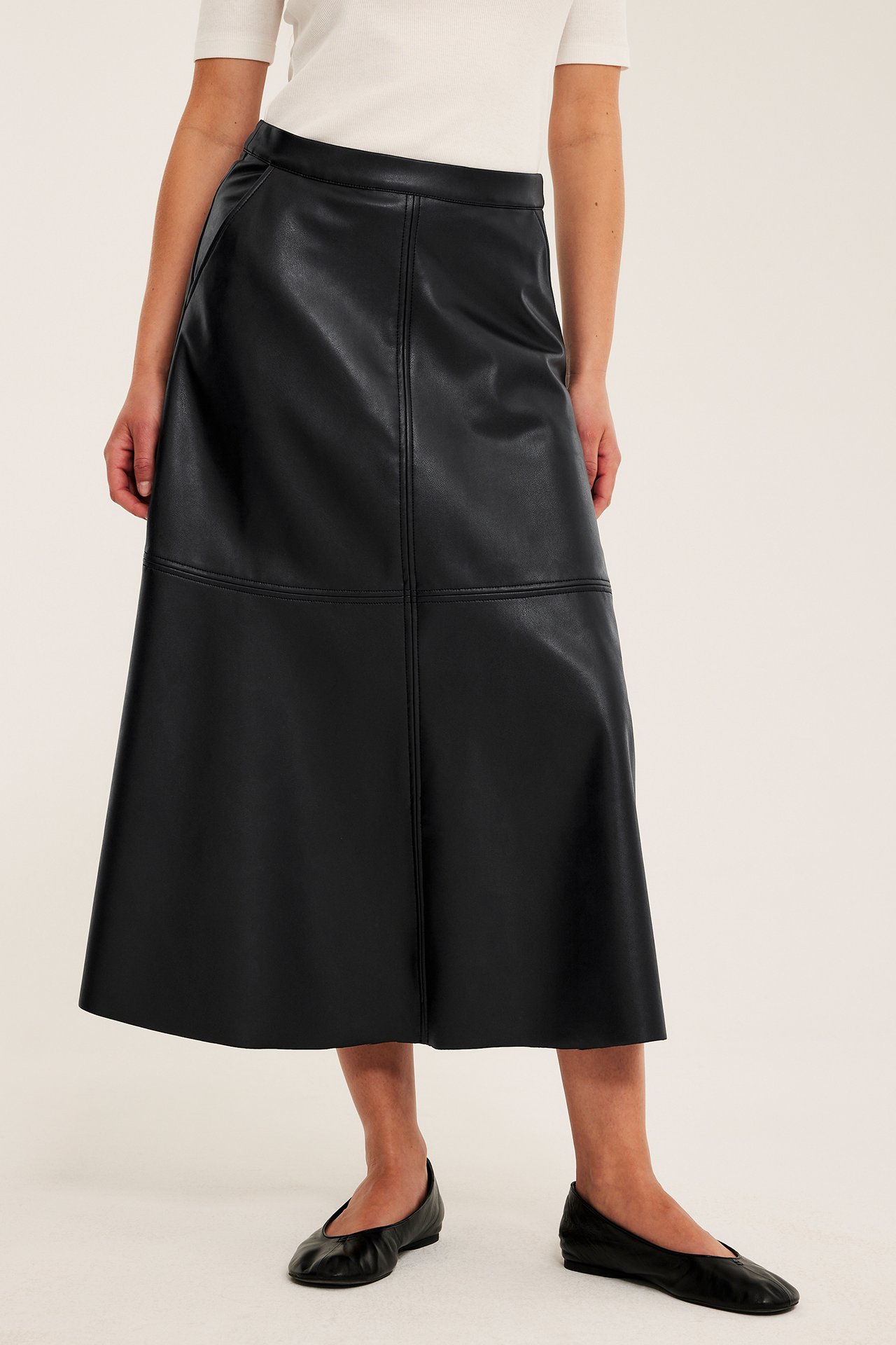 Framifrån: kvinna i svart utsvängd midikjol i skinnimitation, vit t-shirt och svarta ballerinaskor.