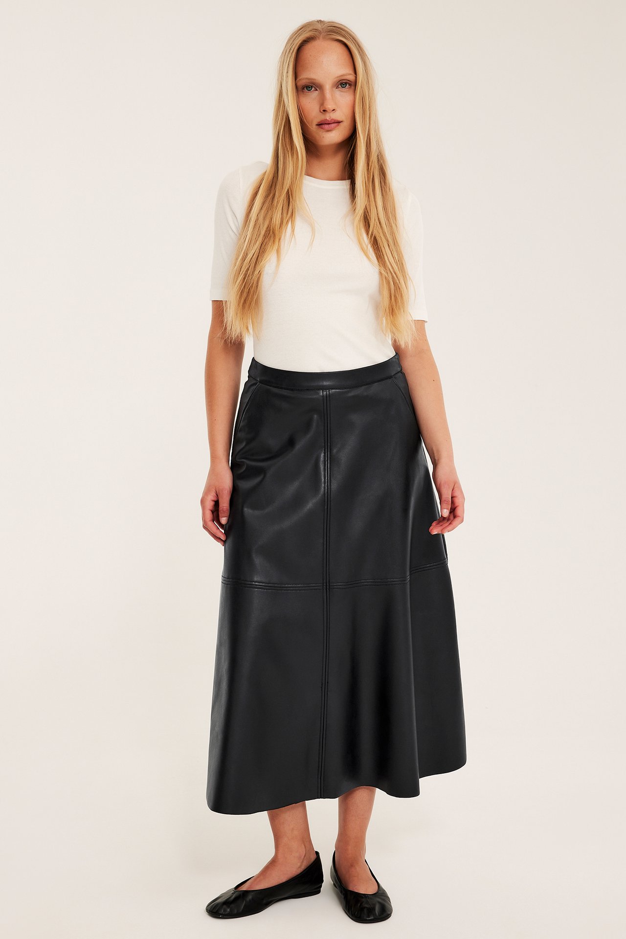 Framifrån: kvinna i svart utsvängd lång kjol i skinnimitation, vit t-shirt och svarta platta skor.