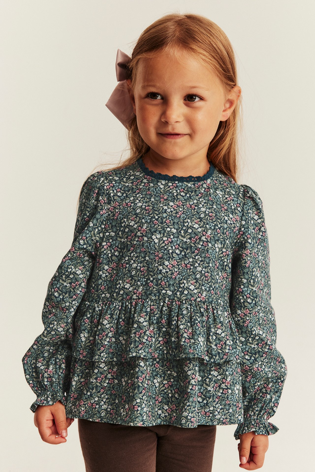Frontvy: Barn i mörkblommig långärmad topp med volanger, bruna leggings och lila hårrosett.