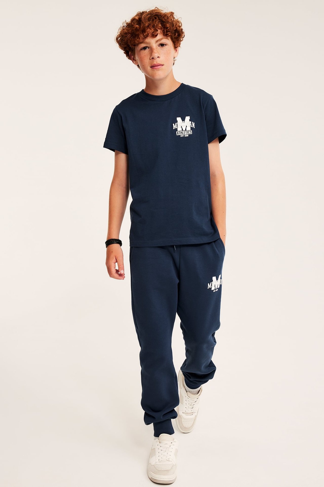 Framifrån, barn i mörkblå tryckt t-shirt, mjukisbyxor, vita sneakers och armband.