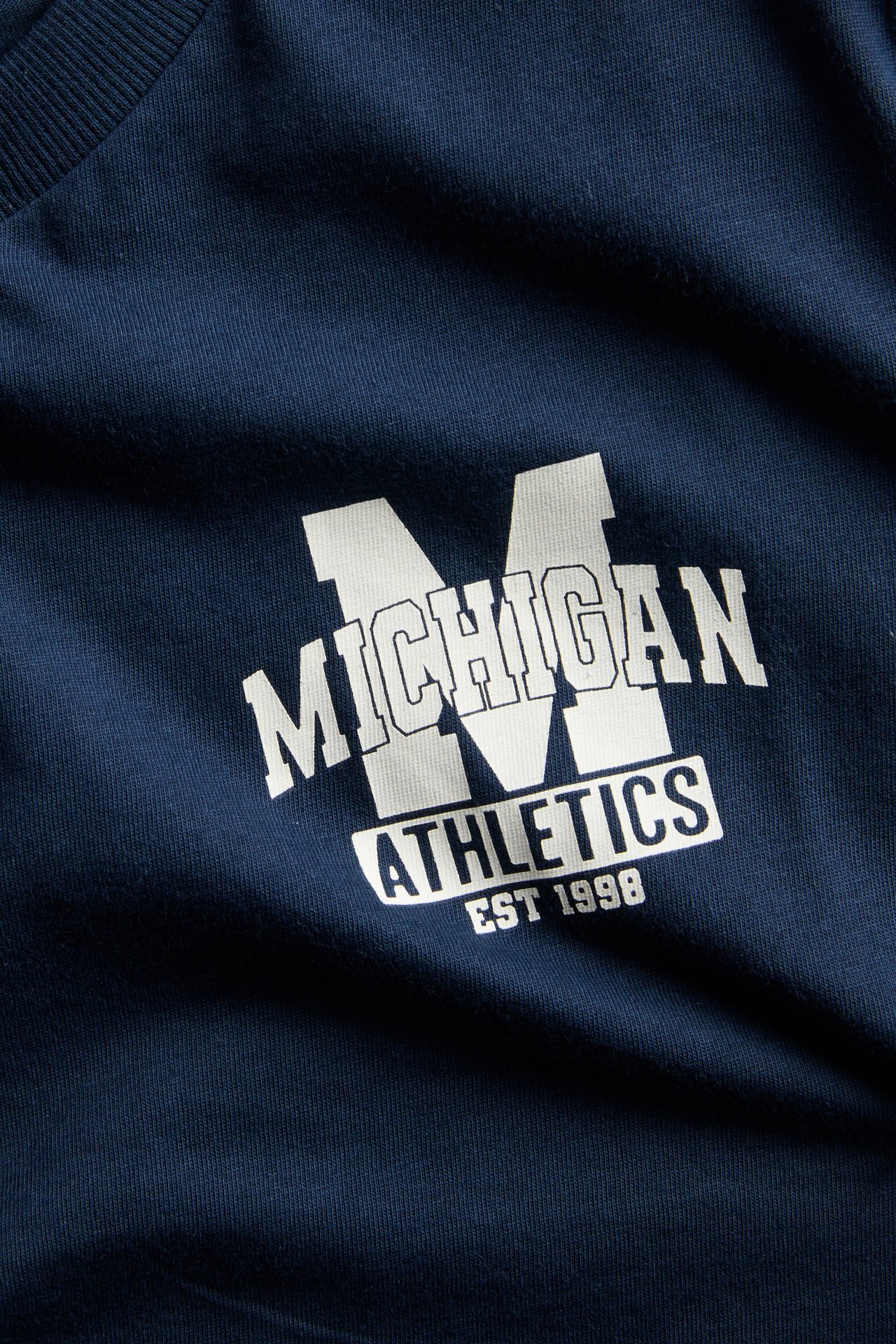 Zbliżenie granatowego t-shirtu z białym nadrukiem „MICHIGAN ATHLETICS EST 1998”.