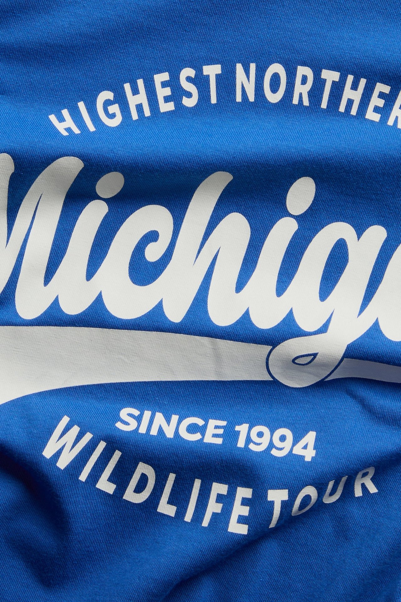 Zbliżenie niebieskiej odzieży z białym nadrukiem: "HIGHEST NORTHERN Michigan SINCE 1994 WILDLIFE TOUR".