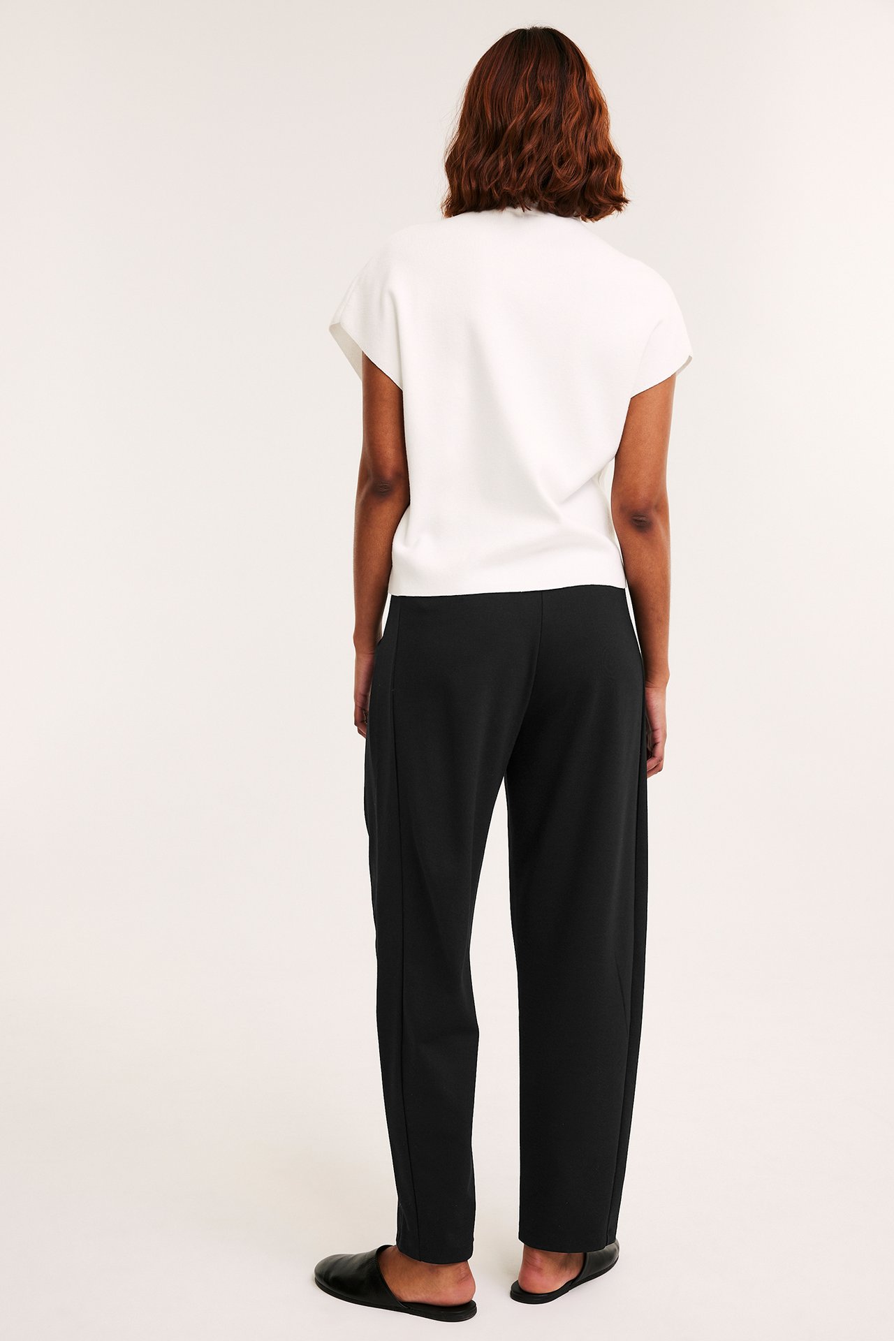 Bakfra: kvinne i svarte barrel leg jerseybukser, hvit topp og svarte flate sko.