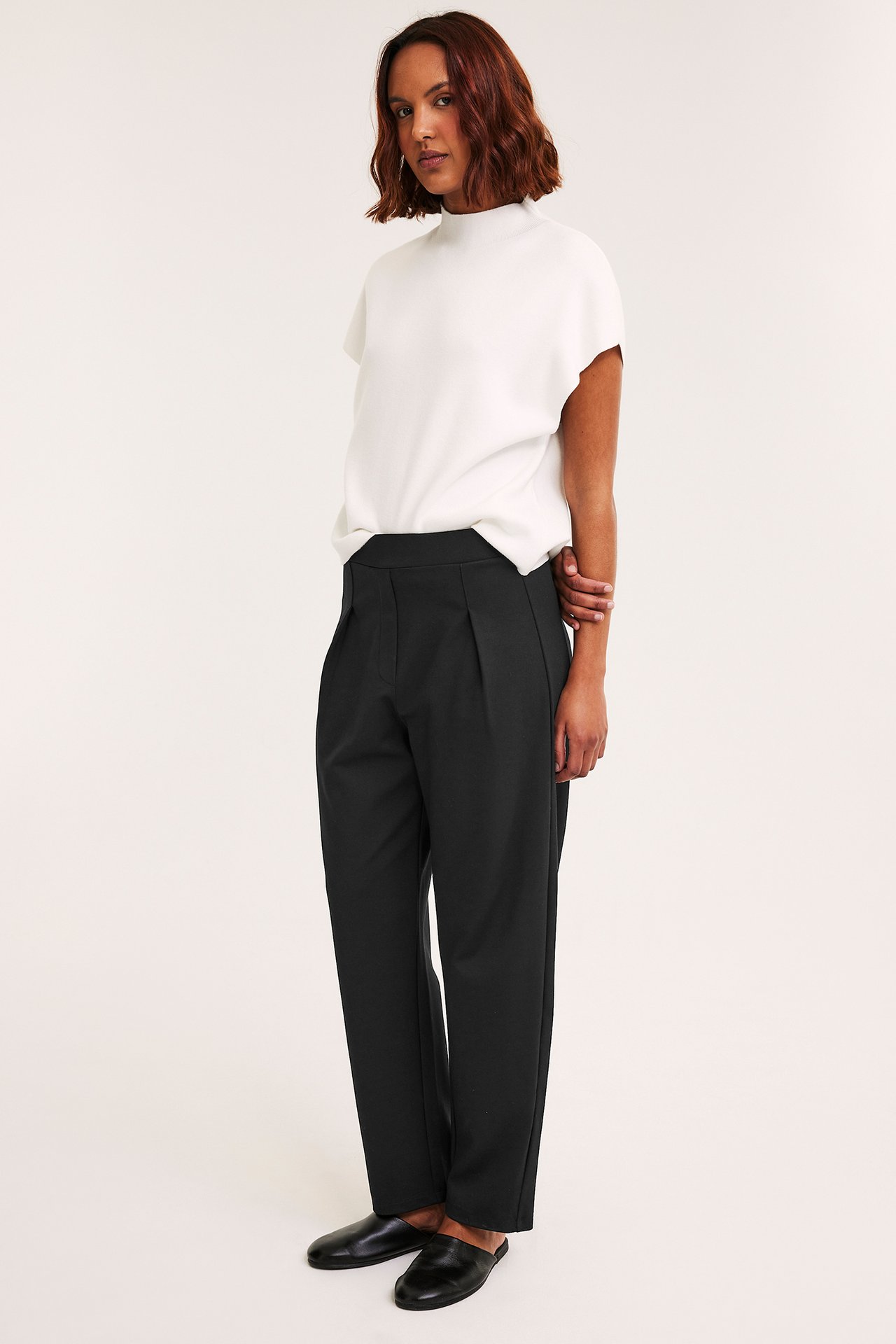 Sivukuva naisesta yllään mustat barrel-lahkeiset trikoohousut, valkoinen poolokauluspaita ja mustat pistokkaat.