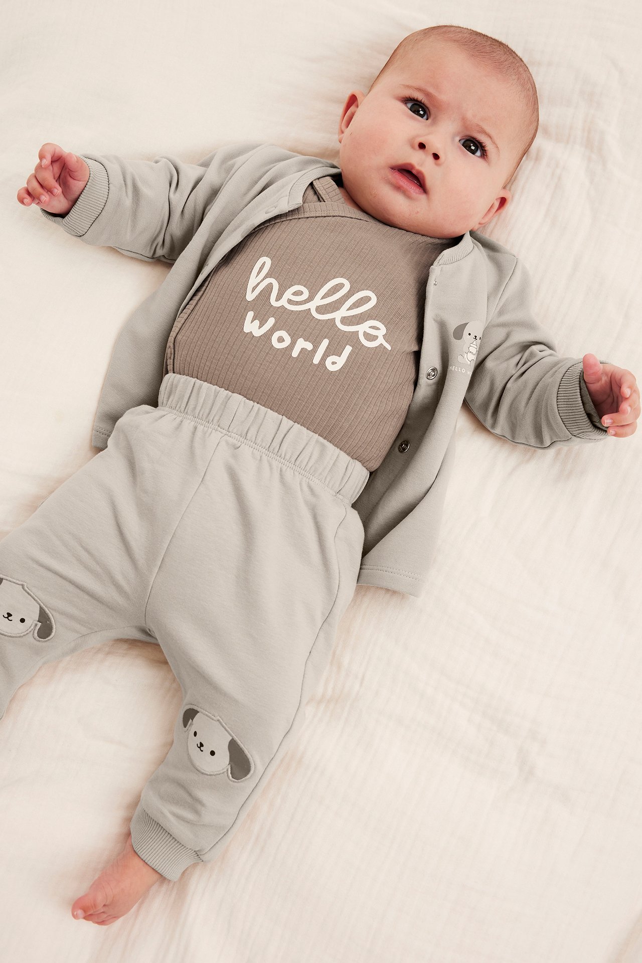 Niemowlak w jasnobrązowym body 'hello world', jasnoszarym kardiganie z psem i pasujących spodniach.