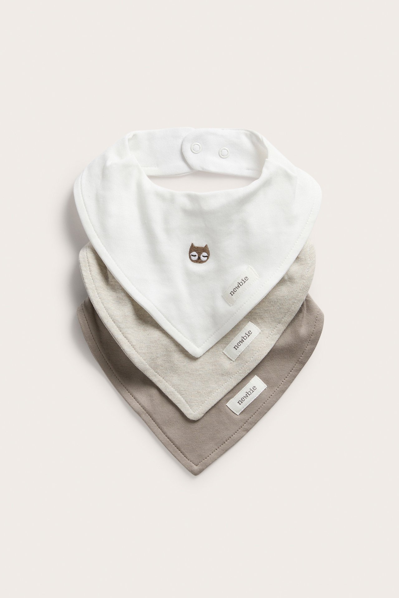 Edestä kolme pinottua vauvan ruokalappua: luonnonvalkoinen pöllöbrodeerauksella, vaalea beige ja taupe.