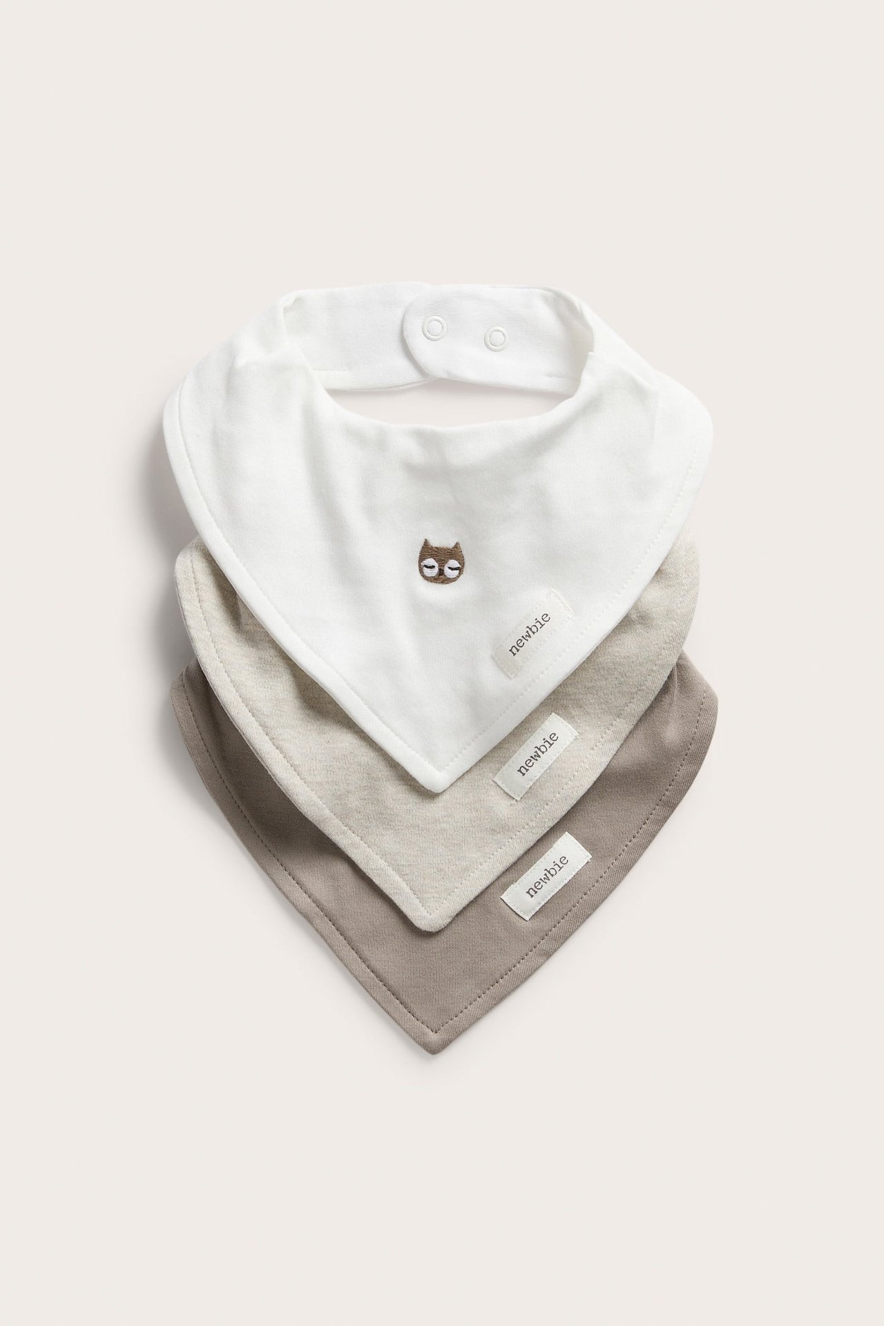 Edestä kolme pinottua vauvan ruokalappua: luonnonvalkoinen pöllöbrodeerauksella, vaalea beige ja taupe.