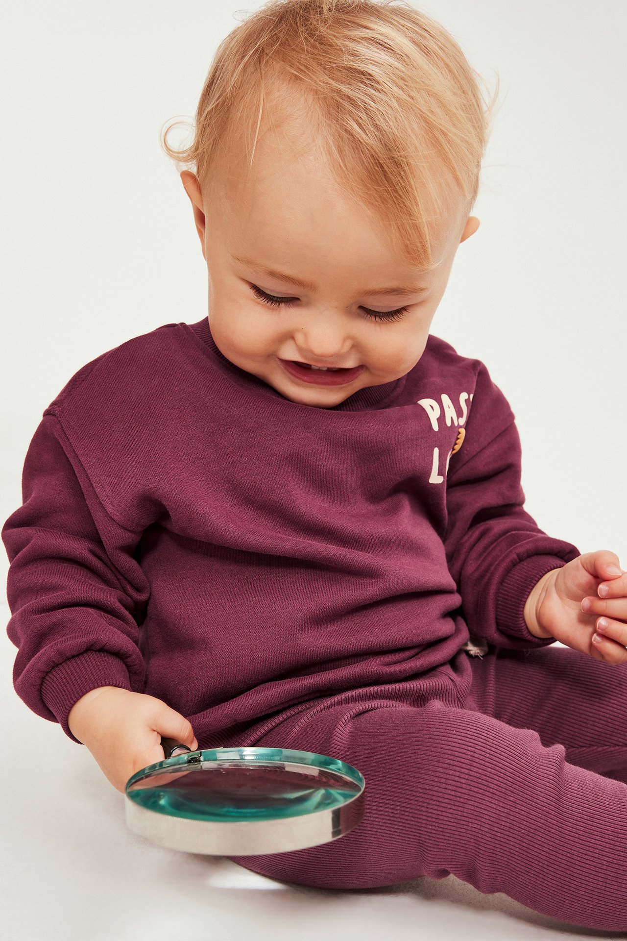 Nærbilde av en baby i mørkerød sweatshirt med delvis trykk og matchende ribbestrikkede leggings, holder forstørrelsesglass.
