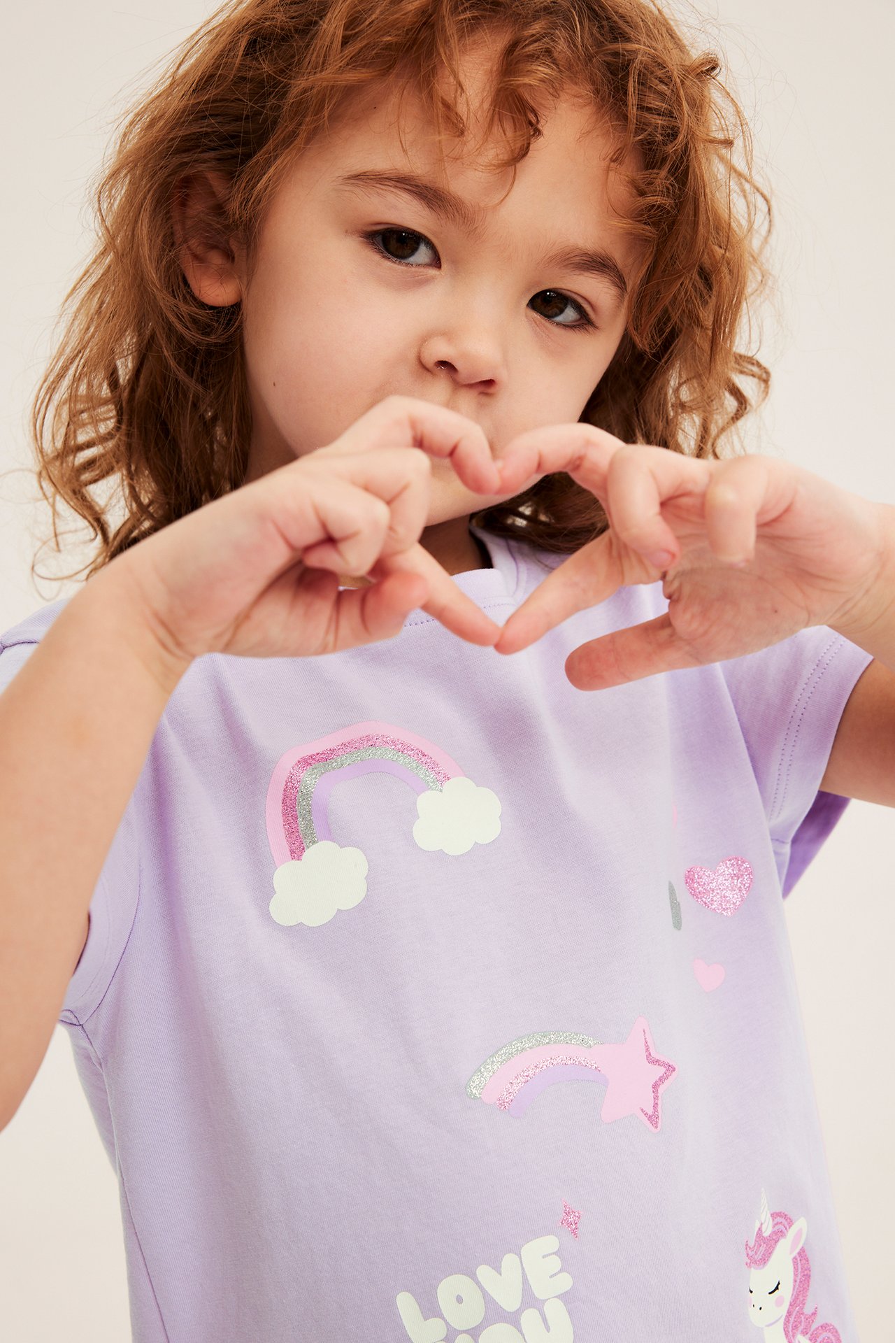 Frontvy: Ett barn i en ljuslila bomullst-shirt med normal passform och tryck av enhörning och regnbåge.