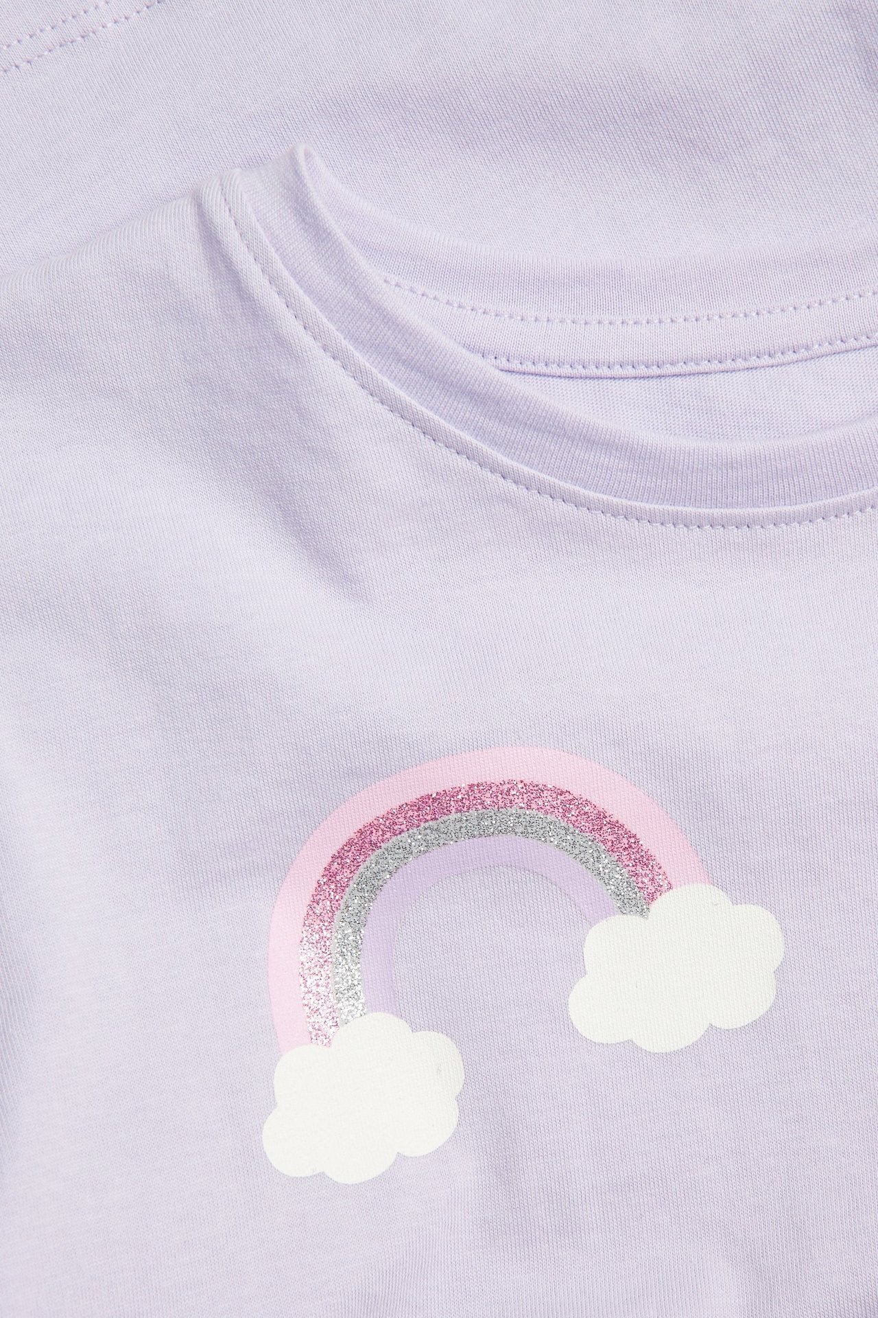 Närbild på en ljuslila barntröja i bomull med ett regnbågstryck i rosa och silverglitter.