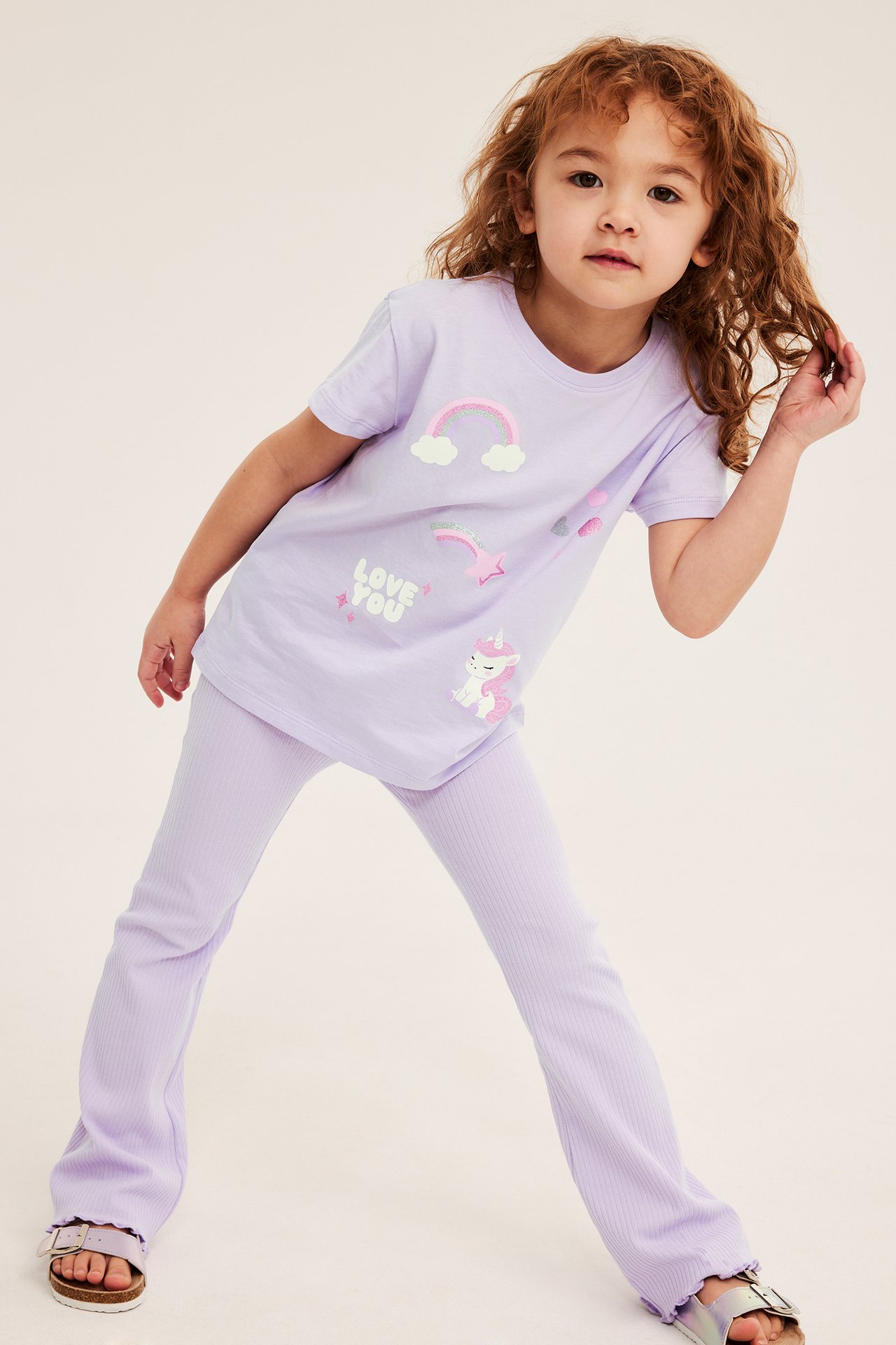 Framifrån, ett barn i ljuslila bomullstopp med enhörningstryck, matchande ribbade leggings och silversandaler.