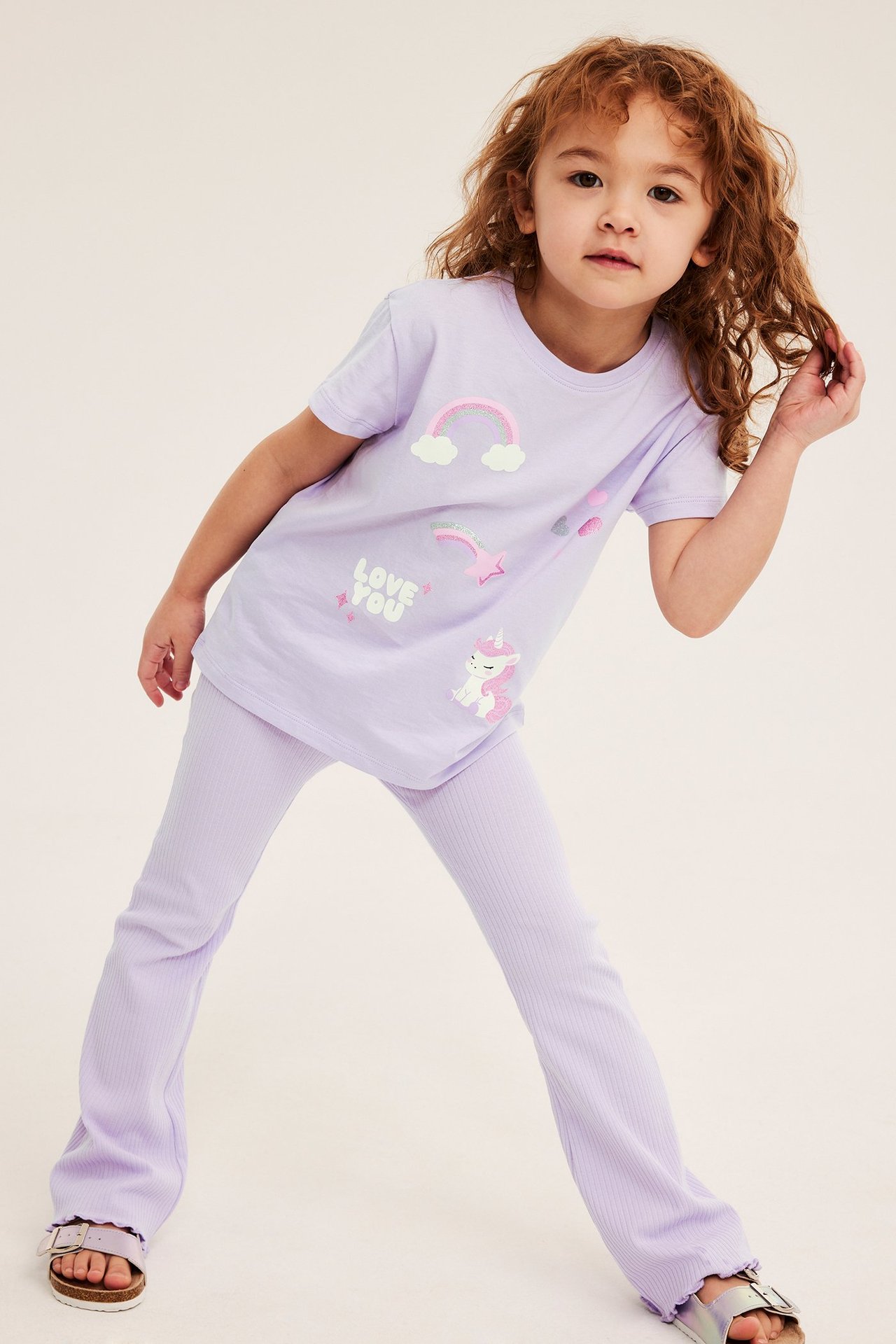 Widok z przodu dziecka w jasnofioletowym bawełnianym T-shircie z nadrukiem jednorożca, prążkowanych legginsach i srebrnych sandałach.