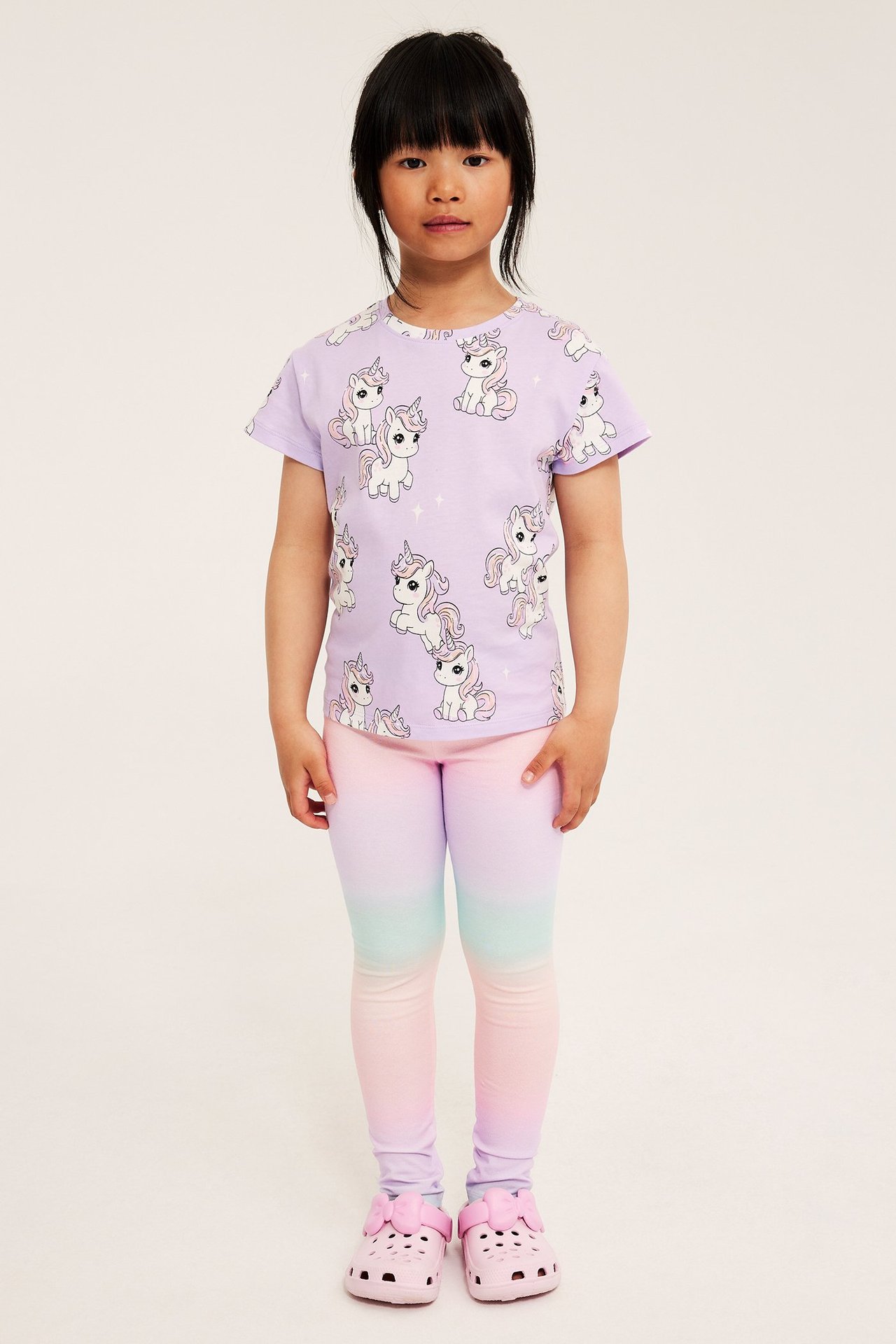 Etunäkymä lapsesta sateenkaarenvärisissä leggingseissä, lilan yksisarvispaidassa ja vaaleanpunaisissa tohveleissa.