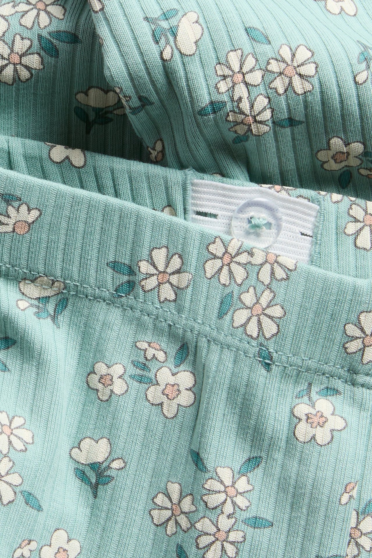 Närbild ljus turkos ribbad barnleggings med vitt blommigt tryck, med justerbar knappmidja.