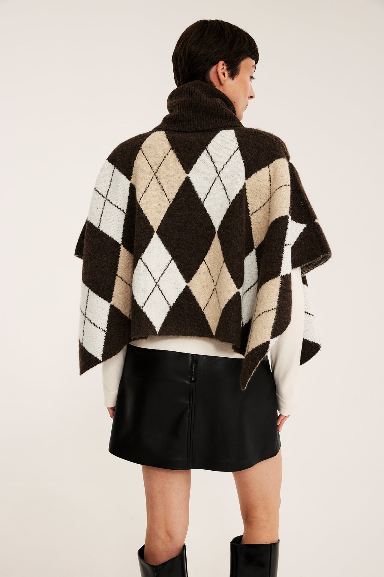 Takaapäin, naismalli yllään ruskea argyle-kuviollinen pooloneuleponcho, valkoinen toppi, musta tekonahkahame ja saappaat.