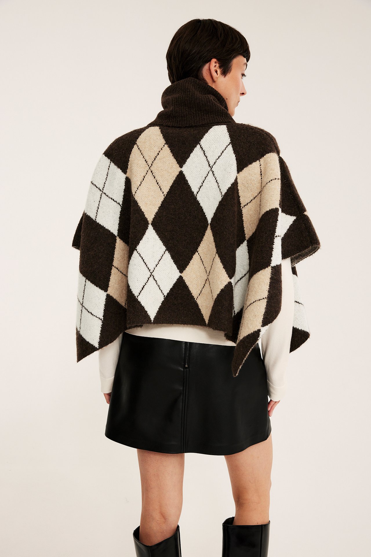 Takaapäin, naismalli yllään ruskea argyle-kuviollinen pooloneuleponcho, valkoinen toppi, musta tekonahkahame ja saappaat.