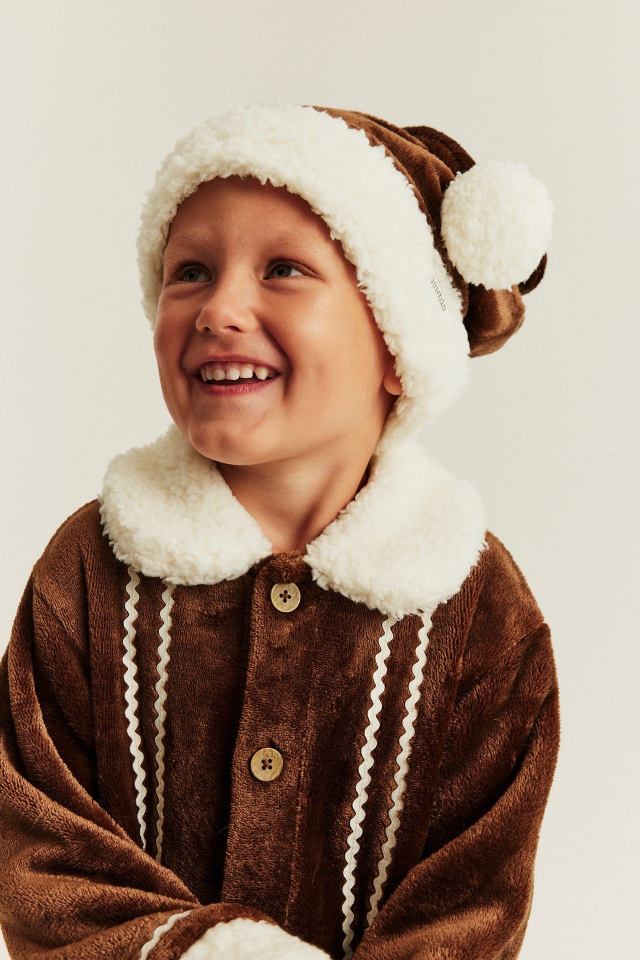 Framifrån på barn i brun plysch pepparkaks-pyjamas set med knappar och mössa.