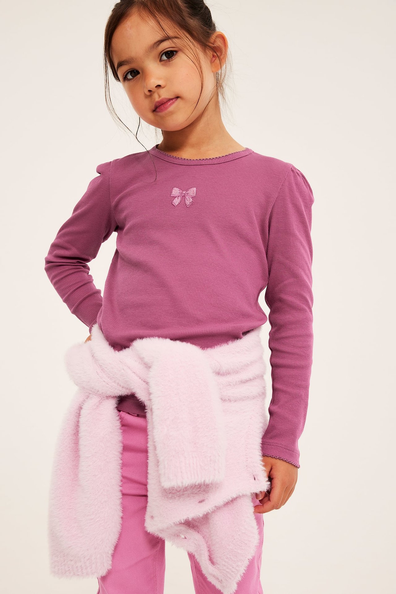 Framsida av flickas lila ribbade långärmade topp med rosett, rosa byxor och knuten fluffig kofta.