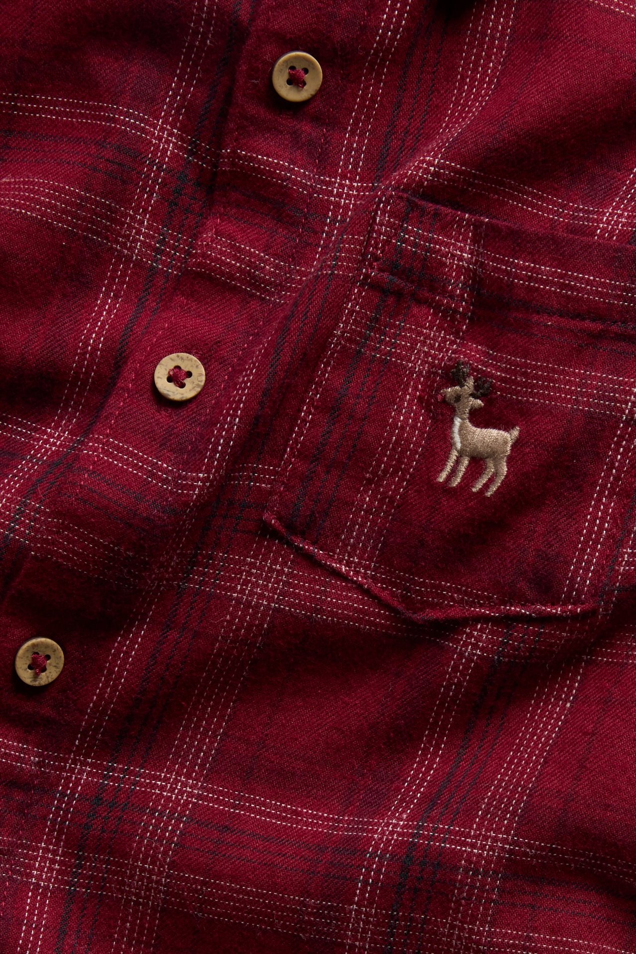 Närbild: Rödrutig flanellskjorta med träknappar och rådjursbroderi på fickan.