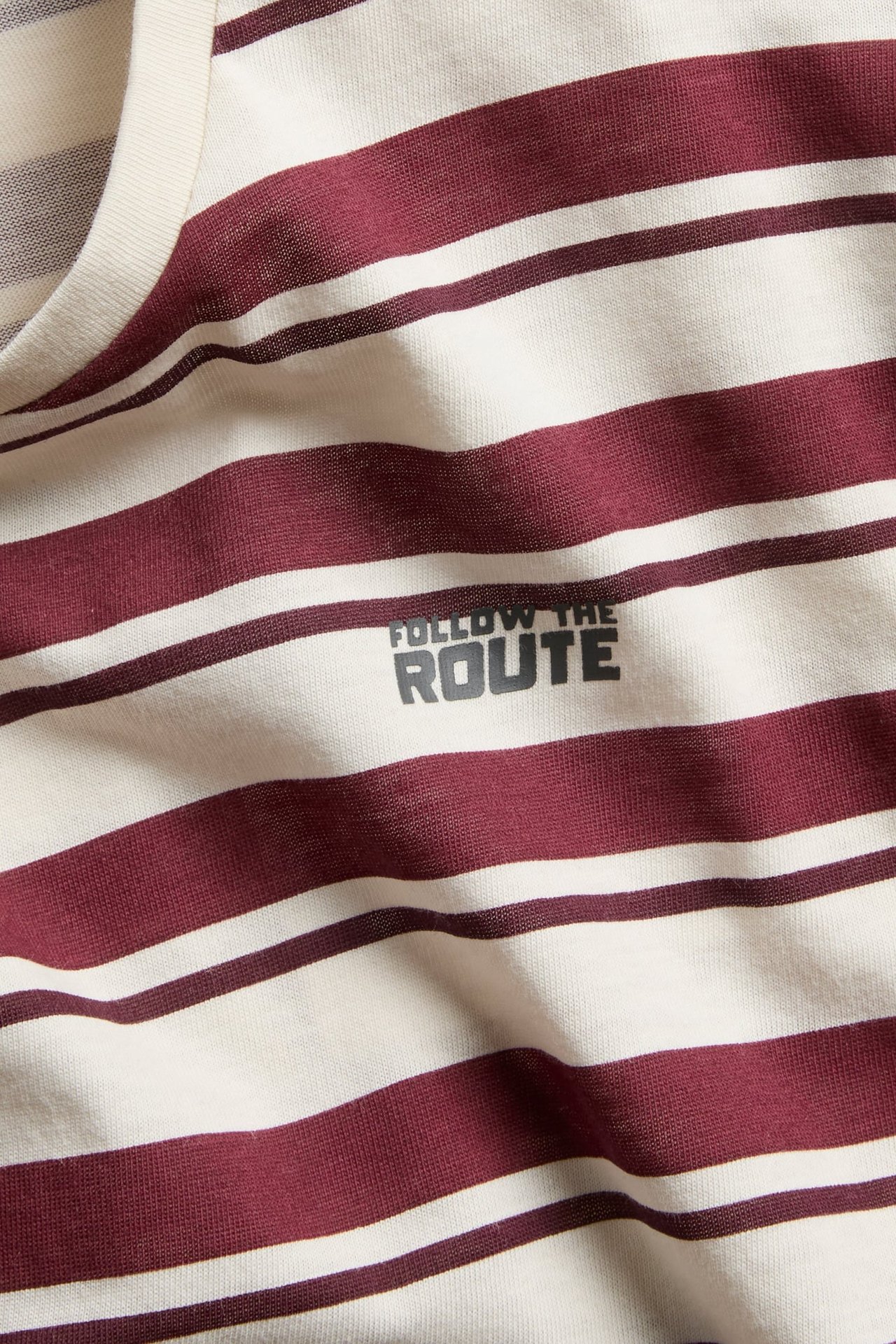 Zbliżenie na pasiastą koszulkę ecru i bordową z nadrukiem 'FOLLOW THE ROUTE'.