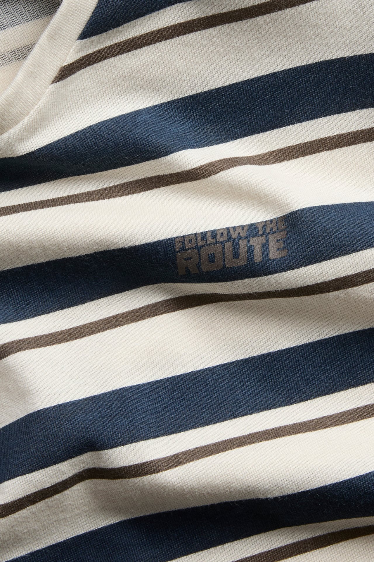 Nærbilde av stripet topp med off-white, mørkeblå og brune horisontale striper, med teksten 'FOLLOW THE ROUTE'.