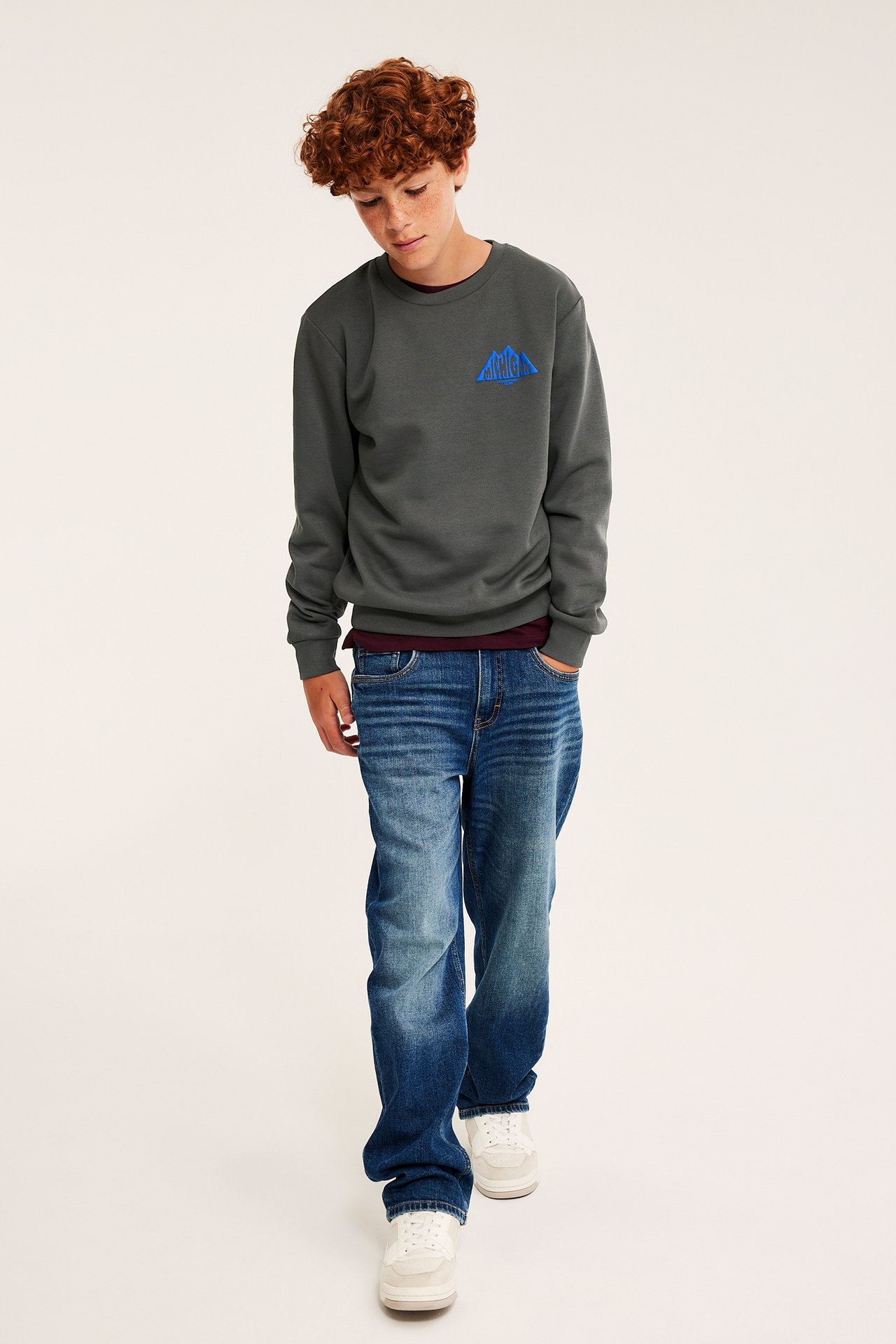 Frontalbilde av barn i mørkegrå langermet genser med blått trykk, blå jeans og hvite joggesko.