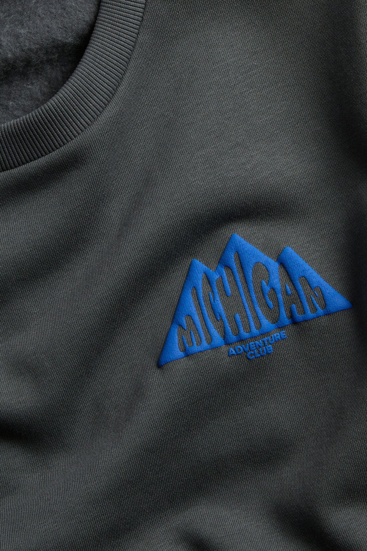 Nærbilde av en mørkegrå crewneck-genser med blått MICHIGAN ADVENTURE CLUB fjellbroderi.