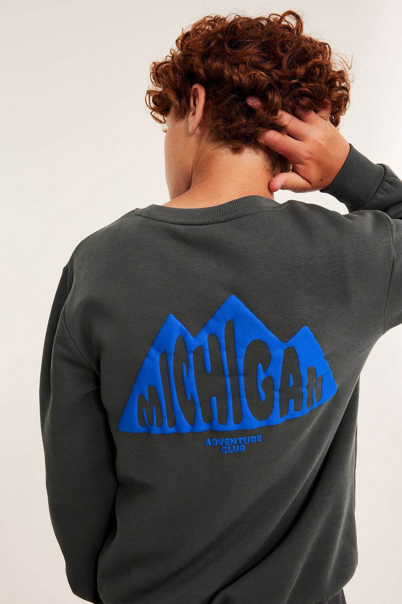 Bakfra av en mannlig modell iført en mørkegrå genser med blå Michigan Adventure Club-grafikk.