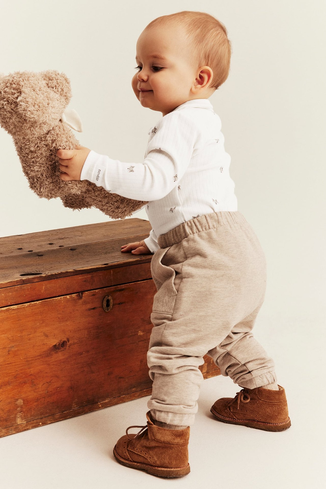 Sidebilde av baby i hvit ribbet mønstret body, beige joggebukse, brune sko, holder en bamse.