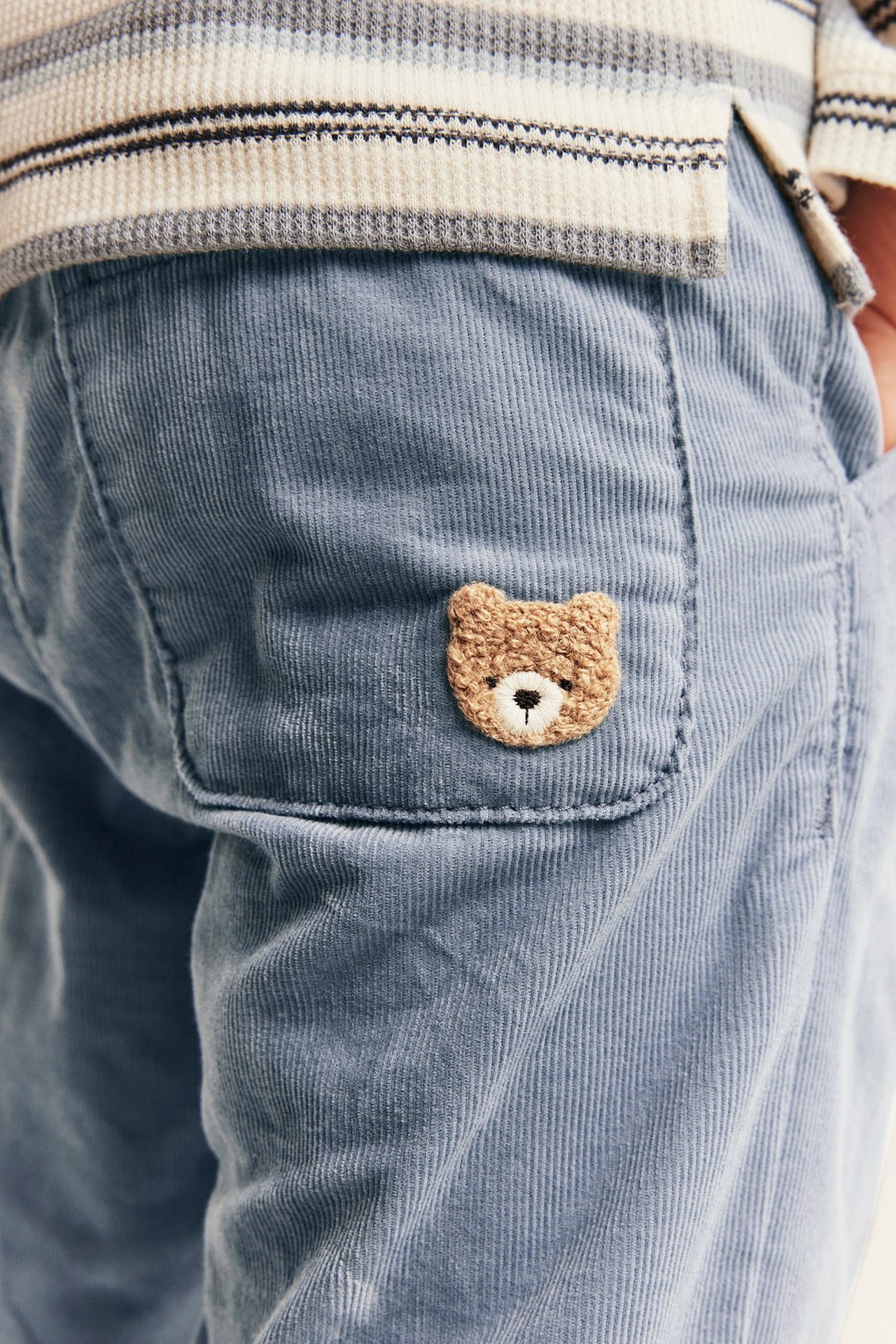 Nærbilde av barnebukse i lyseblå kordfløyel med brodert teddybjørn og stripet topp.