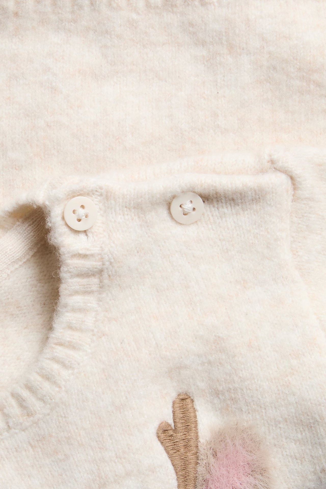 Nærbilde av en kremfarget strikket babygenser med to hvite knapper, brun broderi og rosa lodden detalj.