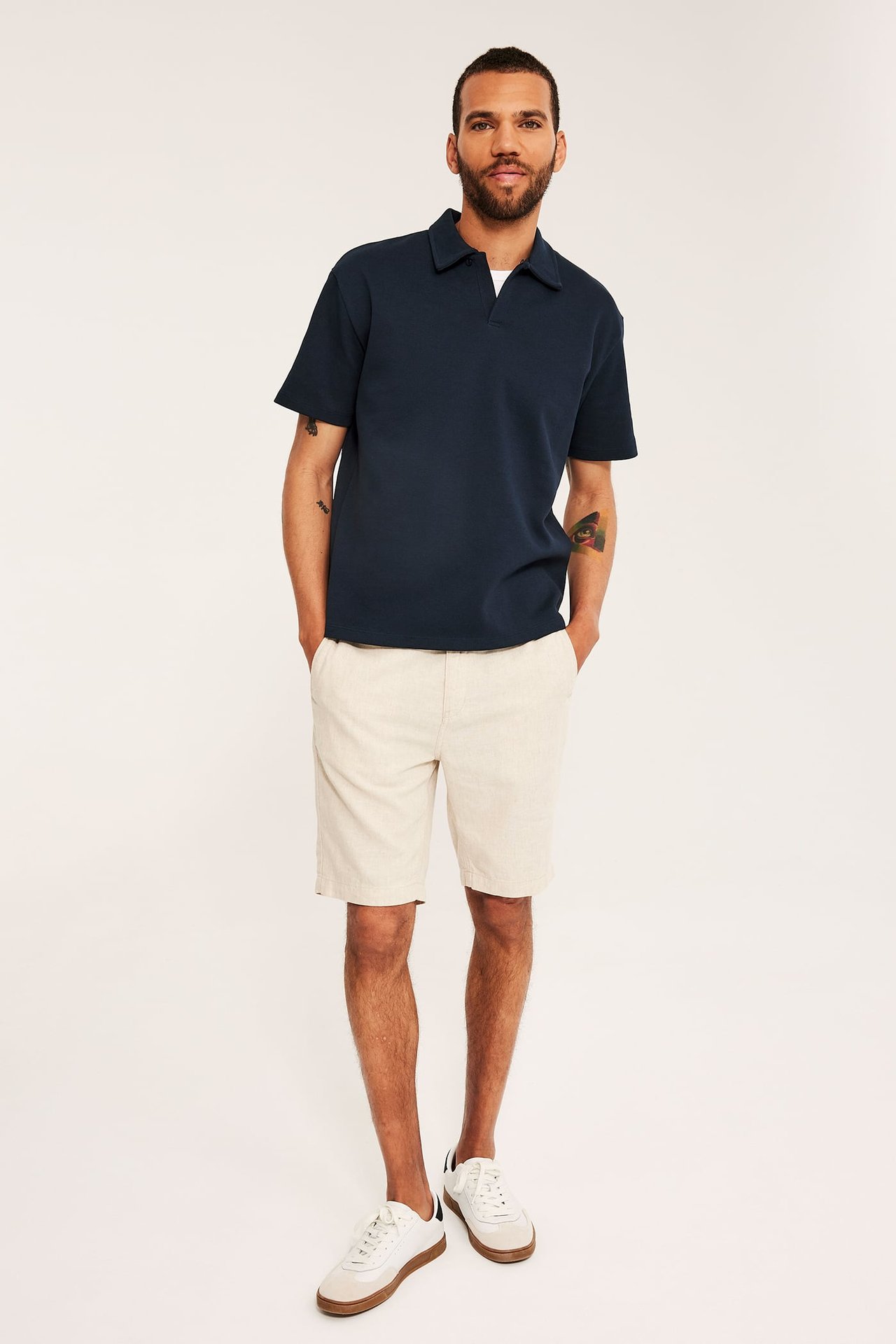 Framifrån: Man i mörkblå lös pikétröja, ljusbeige shorts och vita sneakers.