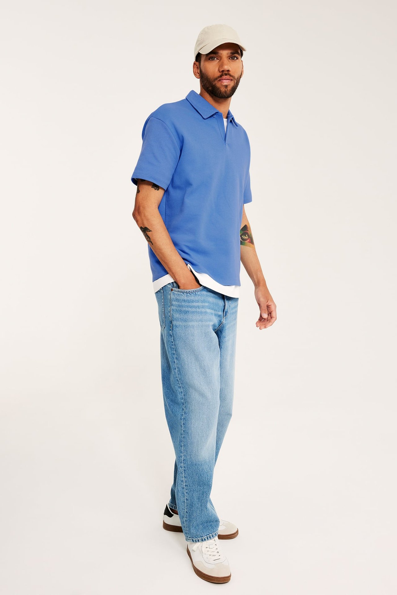 Man i ljusblå lös pikétröja, vit t-shirt, ljusblåa jeans, vita sneakers och beige keps.