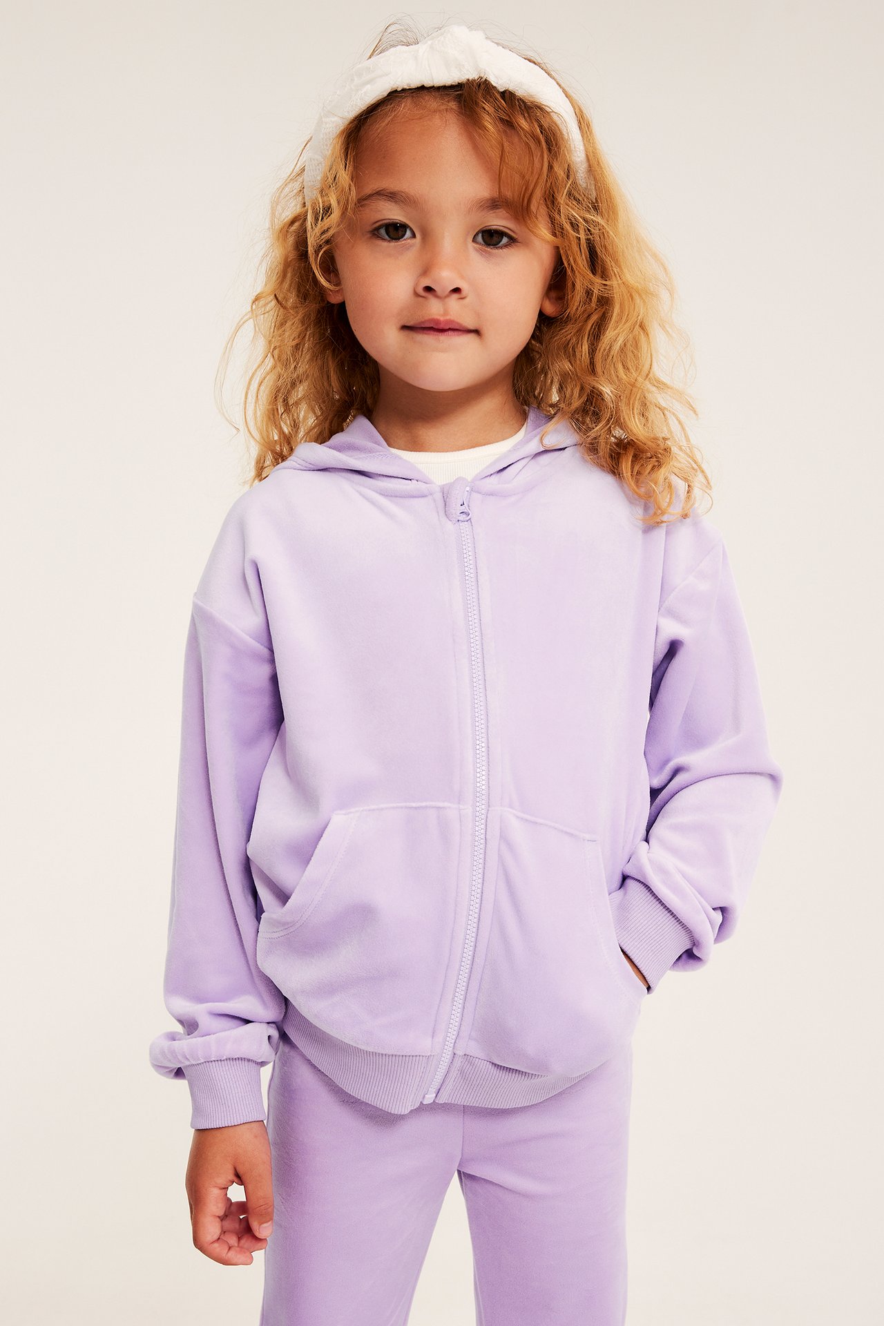Frontvy av ett barn i lila velour huvtröja med dragkedja, matchande sweatpants och vitt knutet diadem.