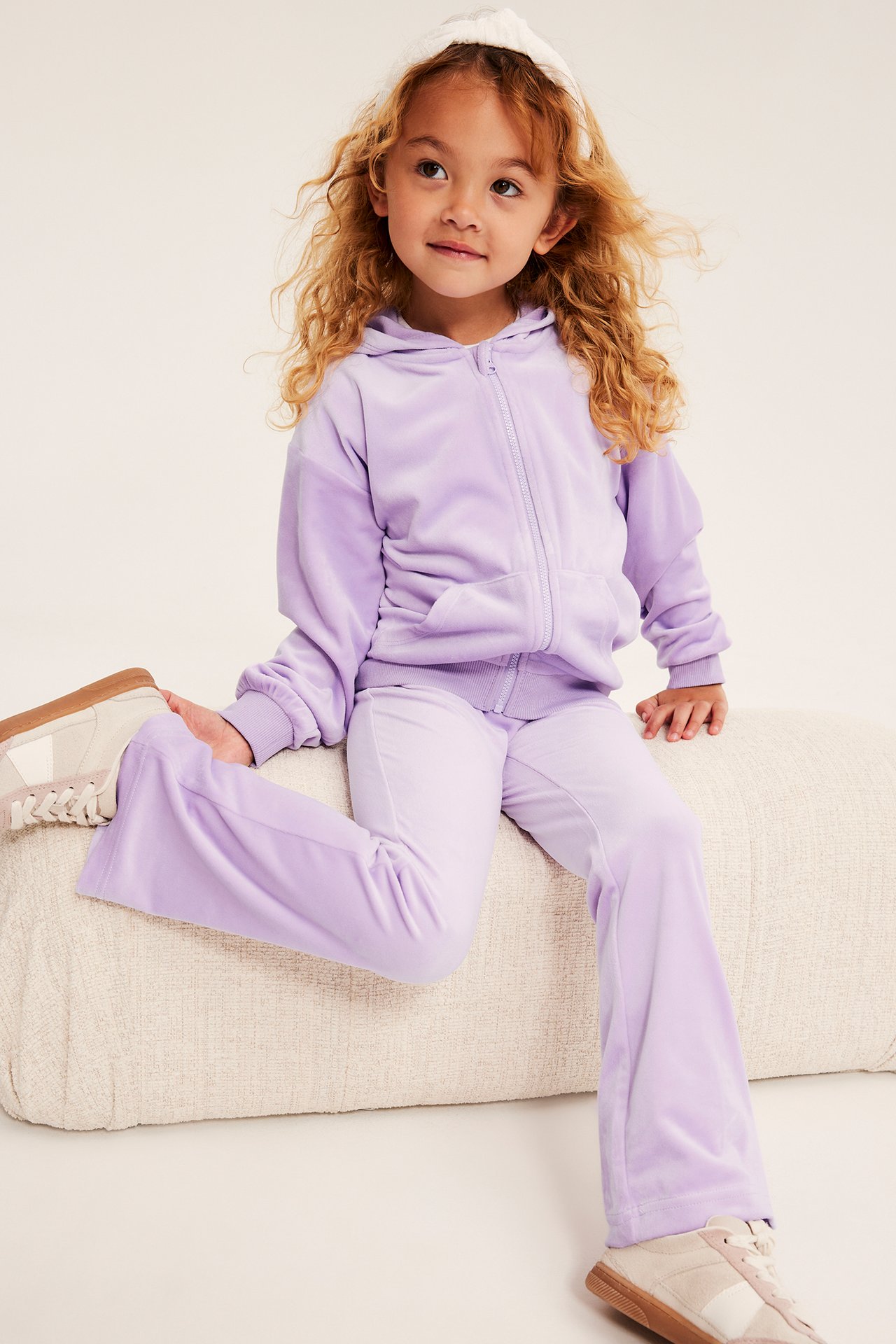 Framsida av ett barn i lila velour tröja med dragkedja och utsvängda byxor med sneakers.