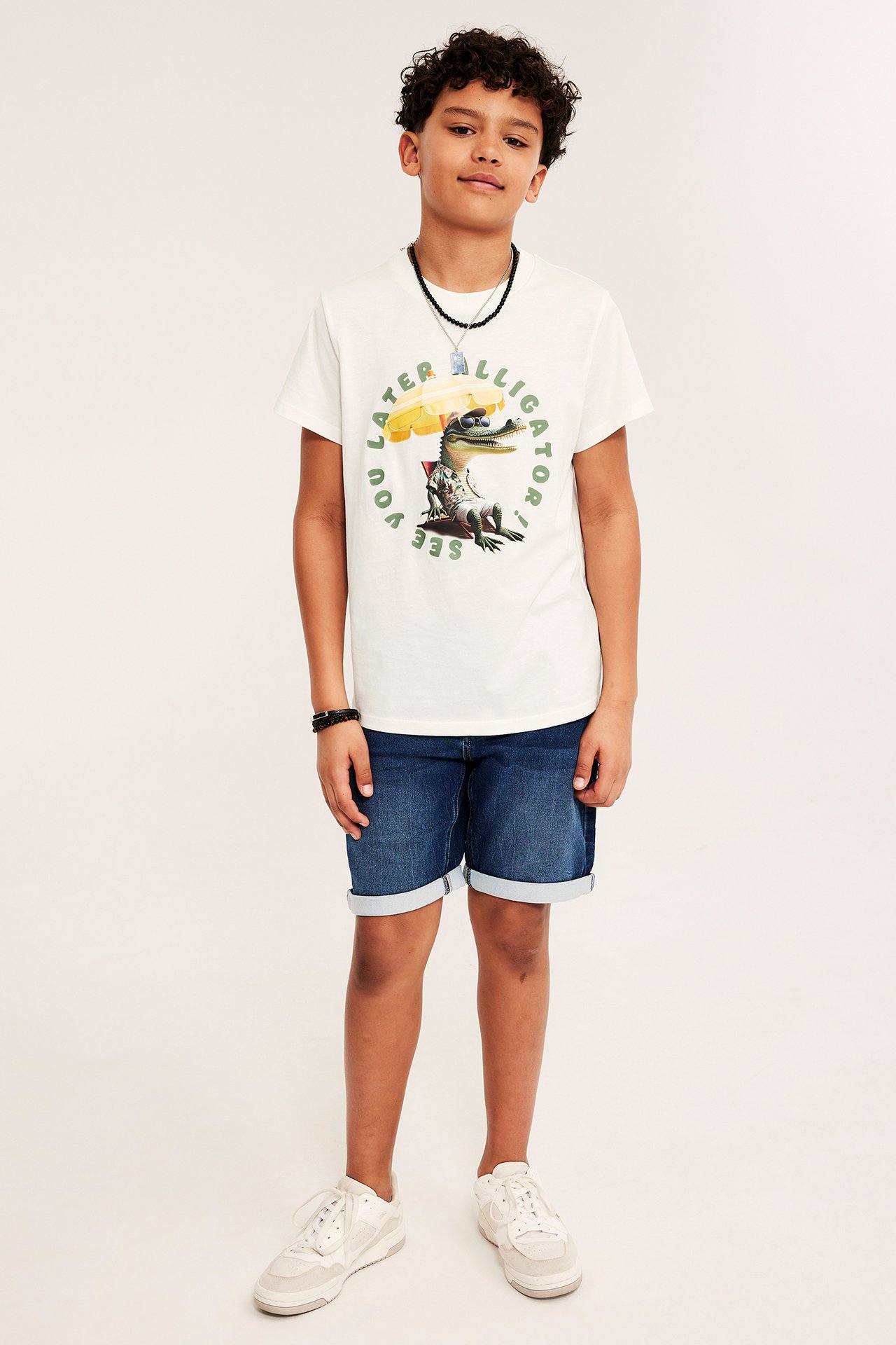 Chłopiec w koszulce bawełnianej off-white z nadrukiem aligatora, niebieskich szortach jeansowych, naszyjniku i białych sneakersach.