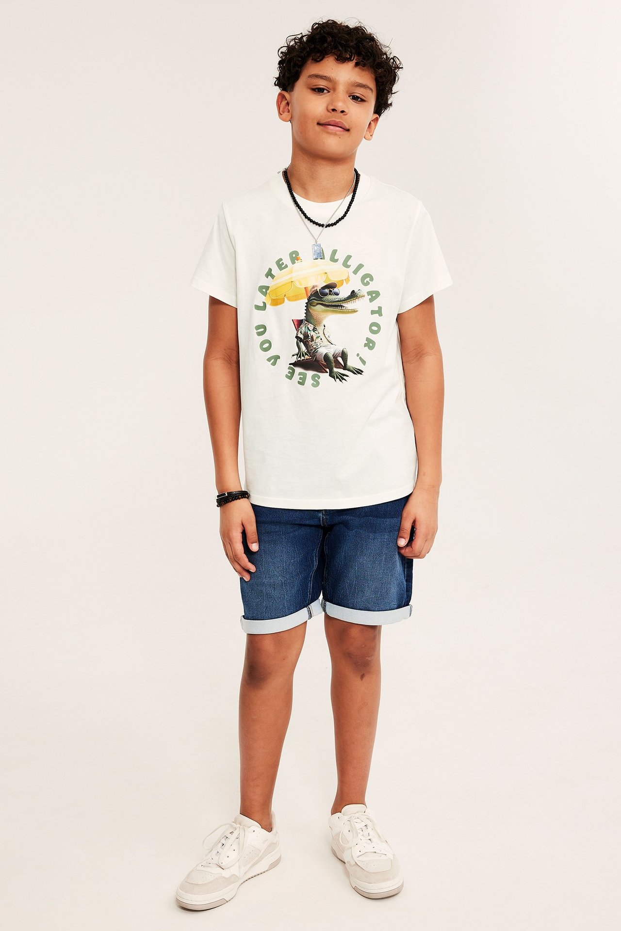 Chłopiec w koszulce bawełnianej off-white z nadrukiem aligatora, niebieskich szortach jeansowych, naszyjniku i białych sneakersach.
