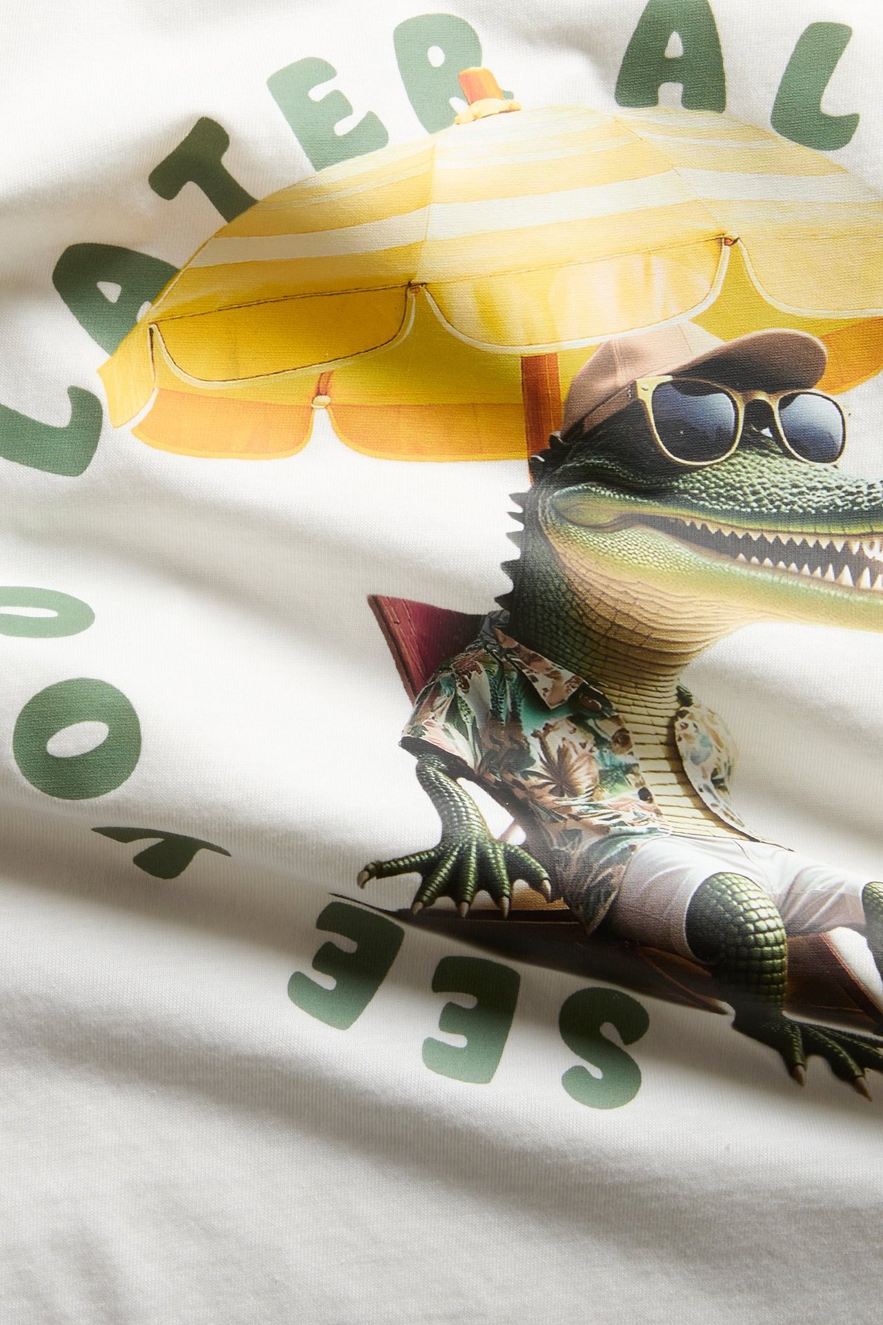 Zbliżenie na bawełniany T-shirt w kolorze ecru z nadrukiem krokodyla w okularach pod parasolem.