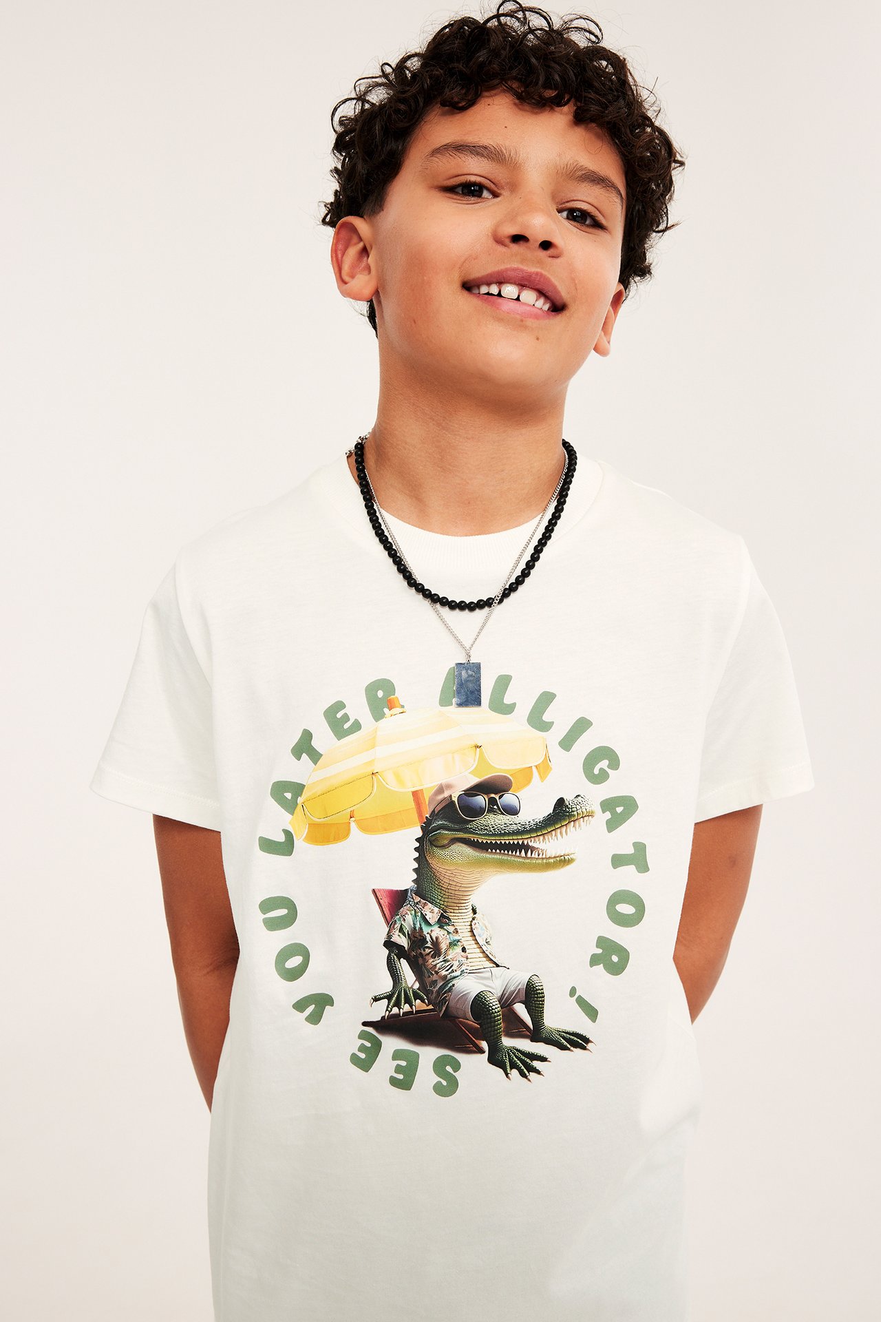 Widok z przodu: chłopiec w kremowym bawełnianym T-shircie z aligatorem i czarnym naszyjniku.