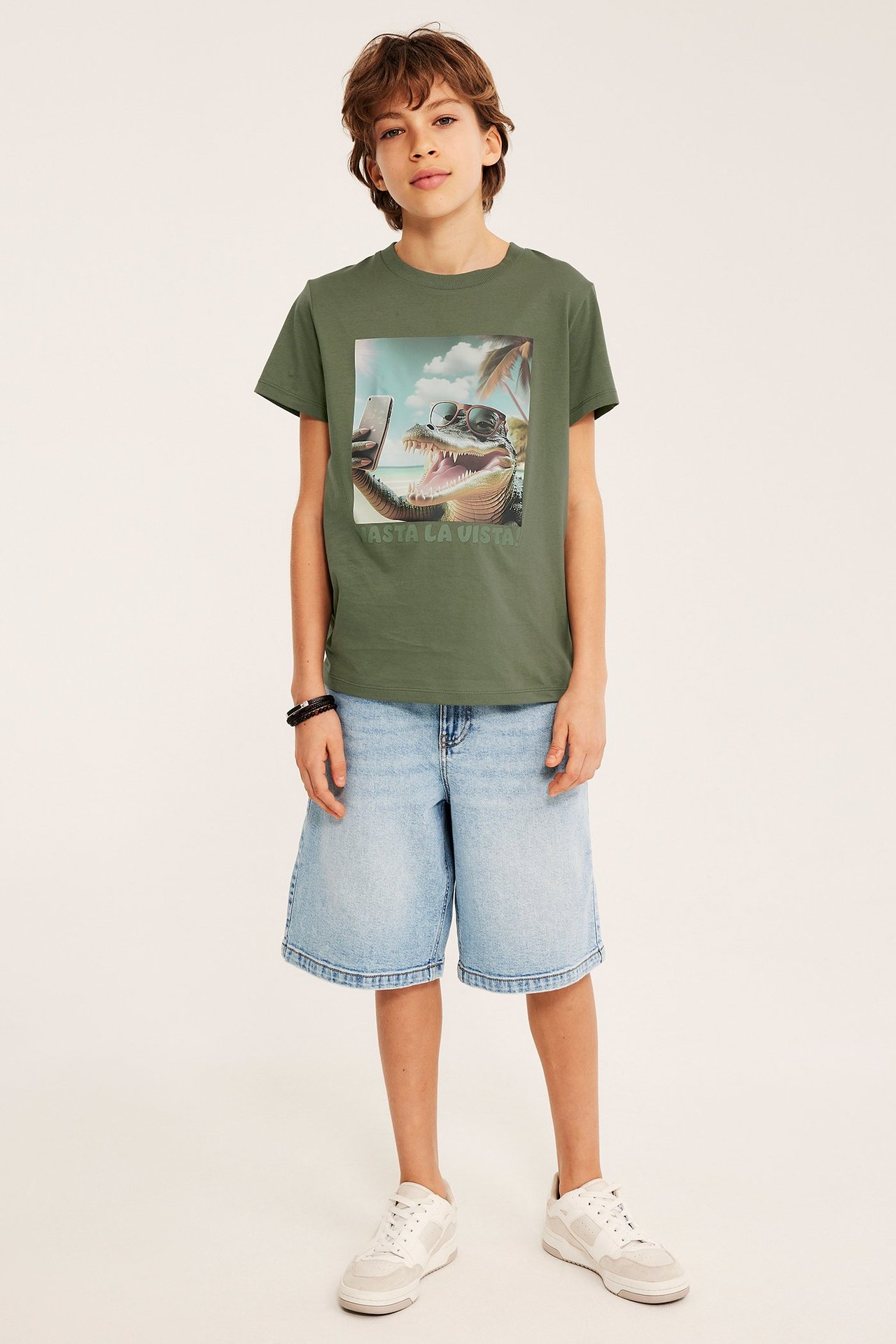 Framifrån: barn i mörkgrön bomulls-T-shirt med krokodiltryck, ljusblå denimshorts och vita sneakers.
