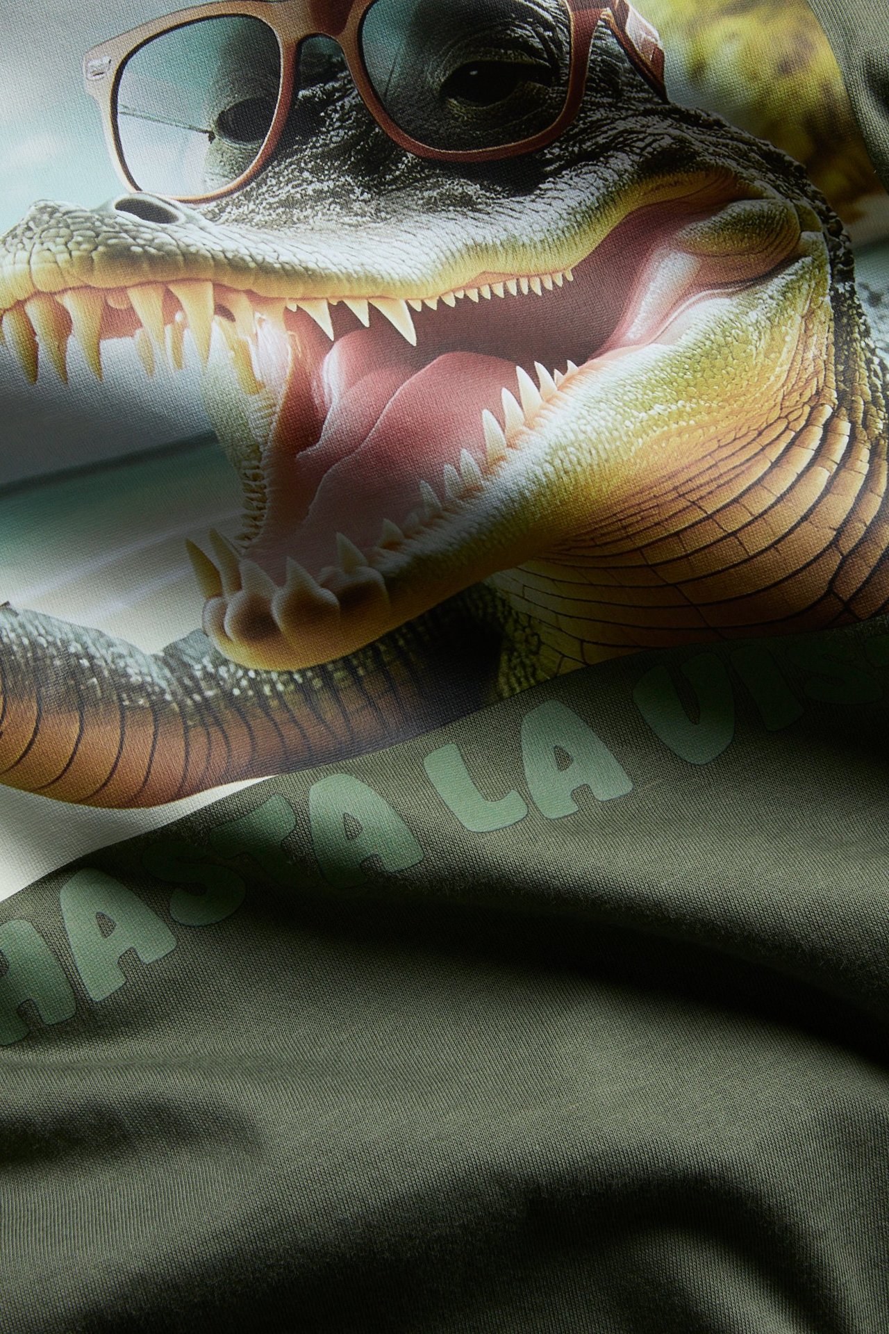 Närbild framifrån: Mörkgrön T-shirt i bomullstrikå med tryck av en alligator i solglasögon.