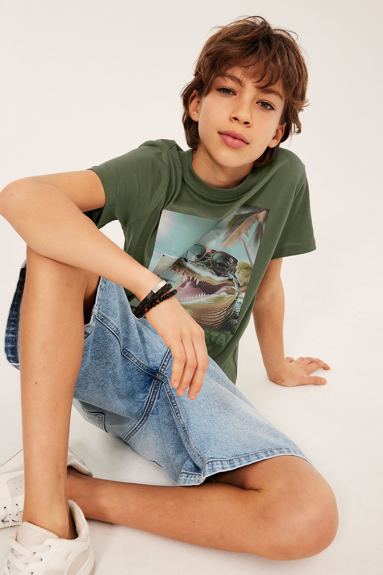 Framifrån: Kid i mörkgrön bomulls-t-shirt med krokodiltryck och ljusblå denimshorts.